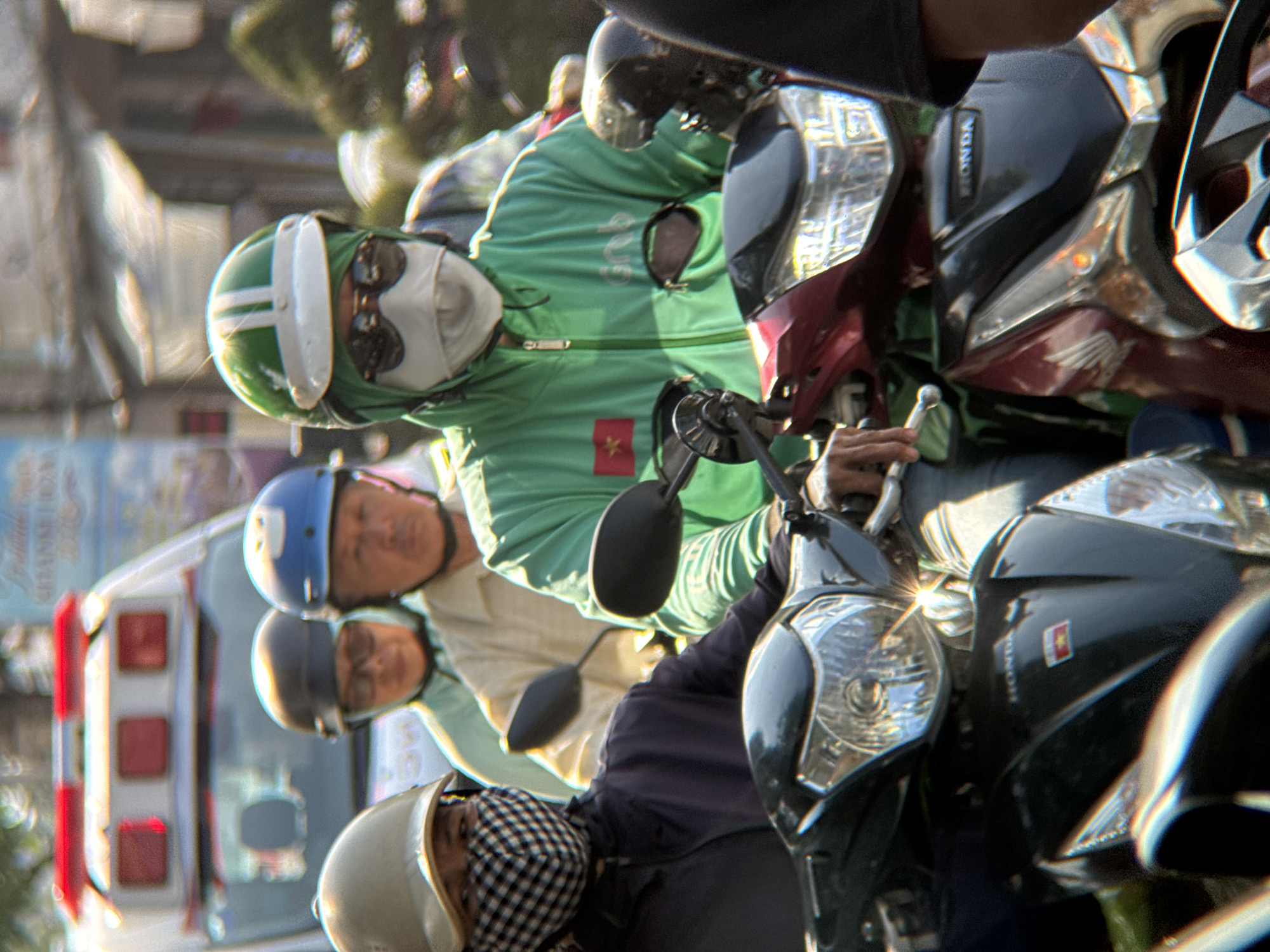 The image shows a group of people on motorcycles, likely in Nha Trang, Vietnam. The foremost person, with a light beige helmet, wears a black and white checkered mask and a dark jacket. Behind them, a person in a light blue helmet and a light yellow shirt is visible. To the right, a person in a green jacket with a white visor and sunglasses is riding a motorcycle. This person has a white mask covering their lower face and a Vietnamese flag emblem on their jacket. The background suggests an urban environment with a large vehicle, possibly an ambulance or service vehicle, and foliage. The motorcycles have visible headlights and handlebars. The lighting indicates it is daytime, with bright sunlight creating highlights and shadows.