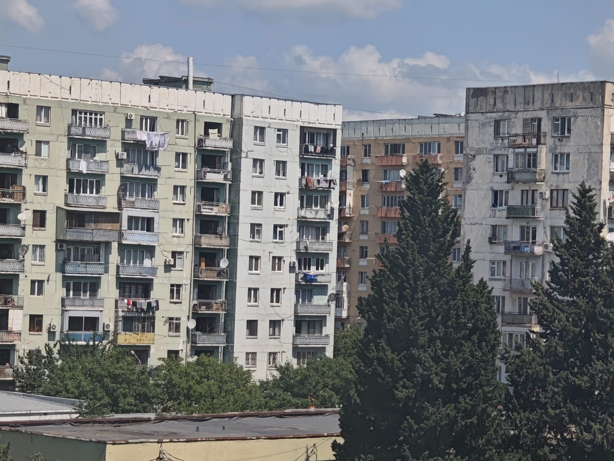 The image shows a view of apartment buildings in the city of Tbilisi, Georgia, likely from a higher vantage point. The buildings are multi-story residential structures with balconies, some of which have clothes drying on them. The architecture suggests a Soviet-era style, common in many parts of the city. Tall, dark green evergreen trees are prominent in the foreground, partially obscuring the lower sections of the buildings and suggesting a residential neighborhood with green spaces. The sky is blue with scattered white clouds, indicating a daytime scene under fair weather conditions. The overall impression is one of an urban residential landscape in Tbilisi.