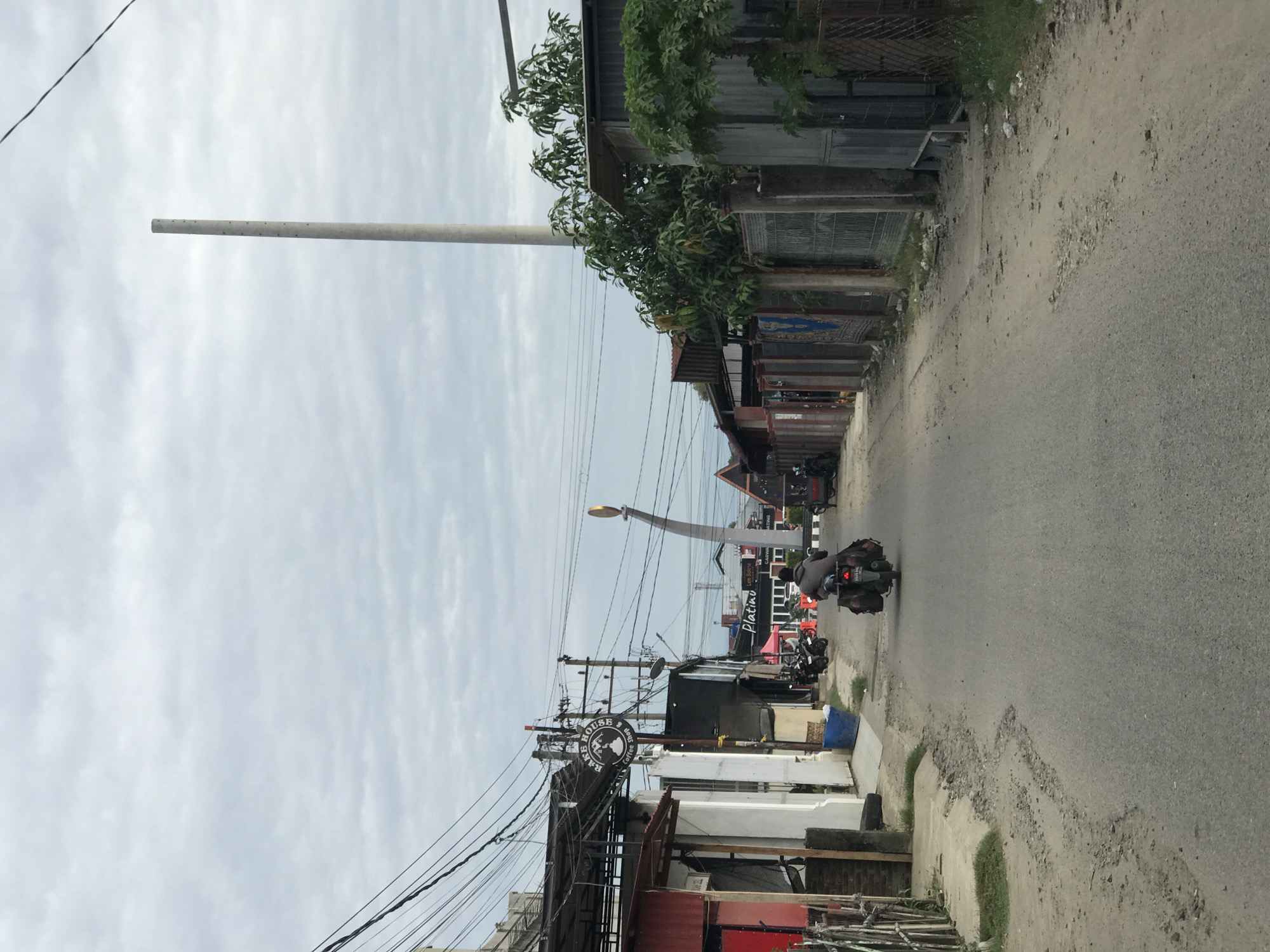 An eye-level, vertical shot captures a street in Lhokseumawe, Indonesia. The street stretches away from the viewer, revealing a narrow road with buildings lining either side. On the right, structures vary from metal corrugated buildings to ones with trees growing on their rooftops. The left side of the street presents a mix of white and red buildings.
Overhead, a web of electrical wires crisscrosses the sky, competing with the cloudy sky. A very tall cylindrical structure stands on the left side of the image. In the distance, a scooter with a rider and luggage is seen travelling toward the viewer. A sign of "Rumoh Rakit Coffee & Shop" is also visible.