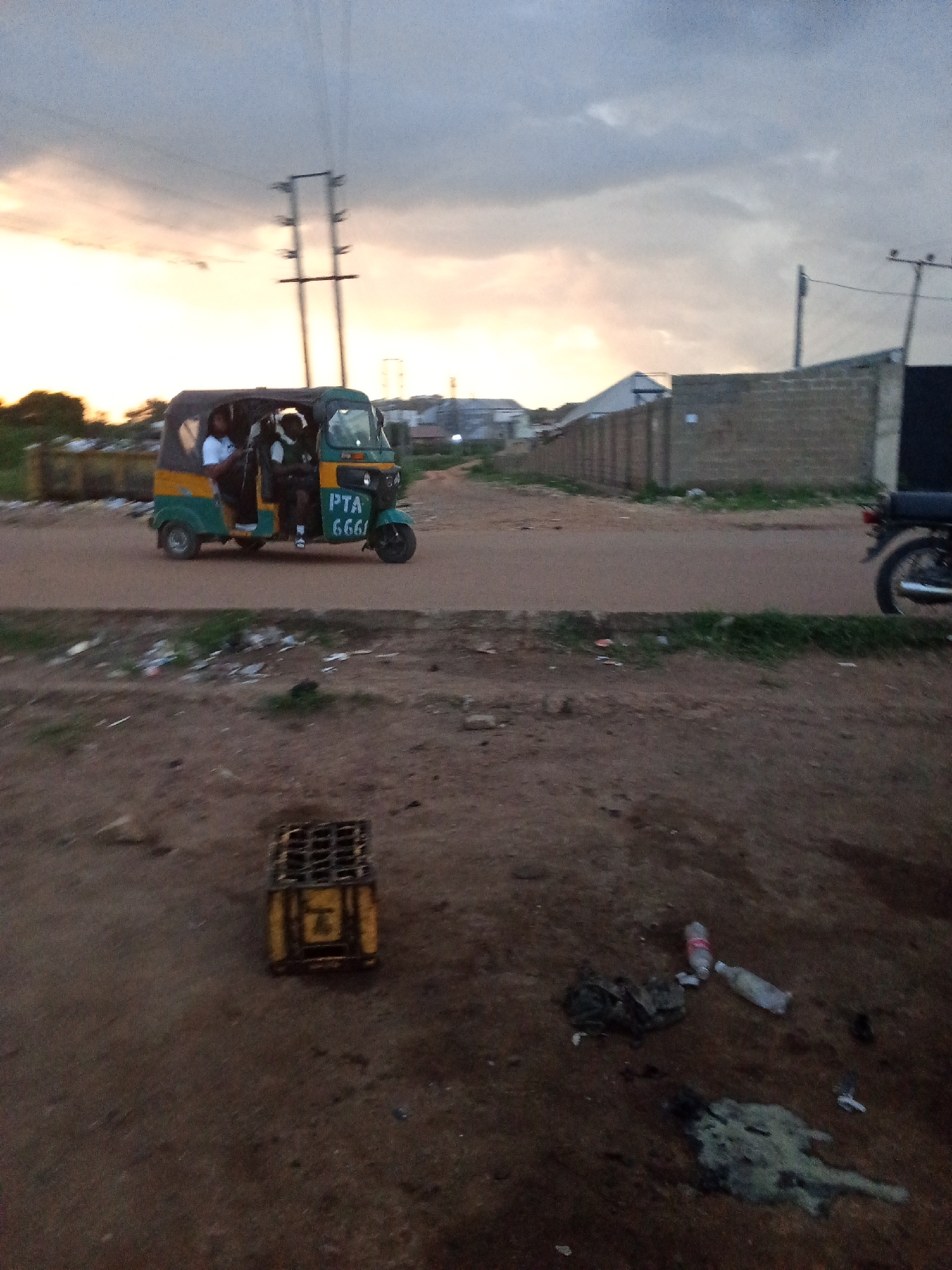 An outdoor, ground-level shot captures a yellow and green auto-rickshaw on a dirt road at dusk, with a cloudy, orange-tinged sky overhead. The auto-rickshaw, driven by a person in a white shirt, carries two passengers. The side of the vehicle displays "PTA 6661" in white lettering. Adjacent to the road, a grey brick wall marks the boundary of a property. In the foreground, a yellow crate with a black grid lid sits on the ground, surrounded by scattered debris, including plastic bottles and dark, amorphous organic matter. A black motorcycle is parked on the right side of the road. The overall scene suggests a somewhat unkempt urban environment in Barakin Lerawi, Nigeria.