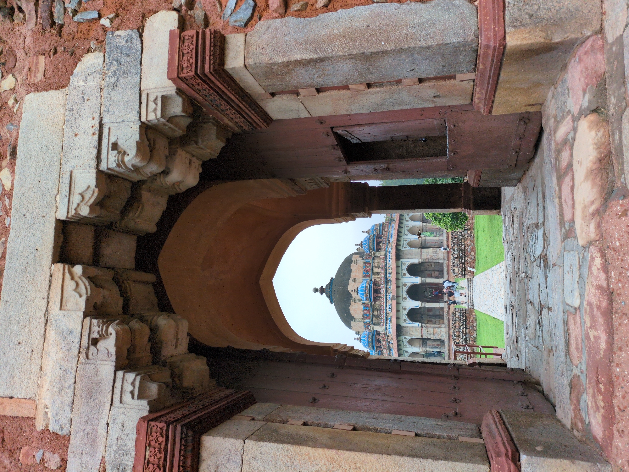 This is a photograph taken from within an ancient stone archway looking out towards a domed building with blue accents. The archway is constructed of large, weathered stone blocks and is flanked by thick, reddish-brown wooden doors, one of which is slightly ajar. Intricate carvings are visible on the stone elements above the archway.
Beyond the archway, a paved pathway leads through a green lawn towards the main building. This building features a large central dome and several smaller domes, all topped with pointed finials. The structure has a colonnaded facade with multiple arched openings. A few people are visible walking on the pathway and near the entrance of the building, suggesting a tourist site.
The architectural style and the presence of a domed mausoleum suggest this location is likely Humayun's Tomb, a UNESCO World Heritage Site in Delhi, India. Humayun's Tomb is renowned for its Mughal architecture and is situated in the Nizamuddin area of Delhi. The overall atmosphere is one of historical grandeur and tranquility.