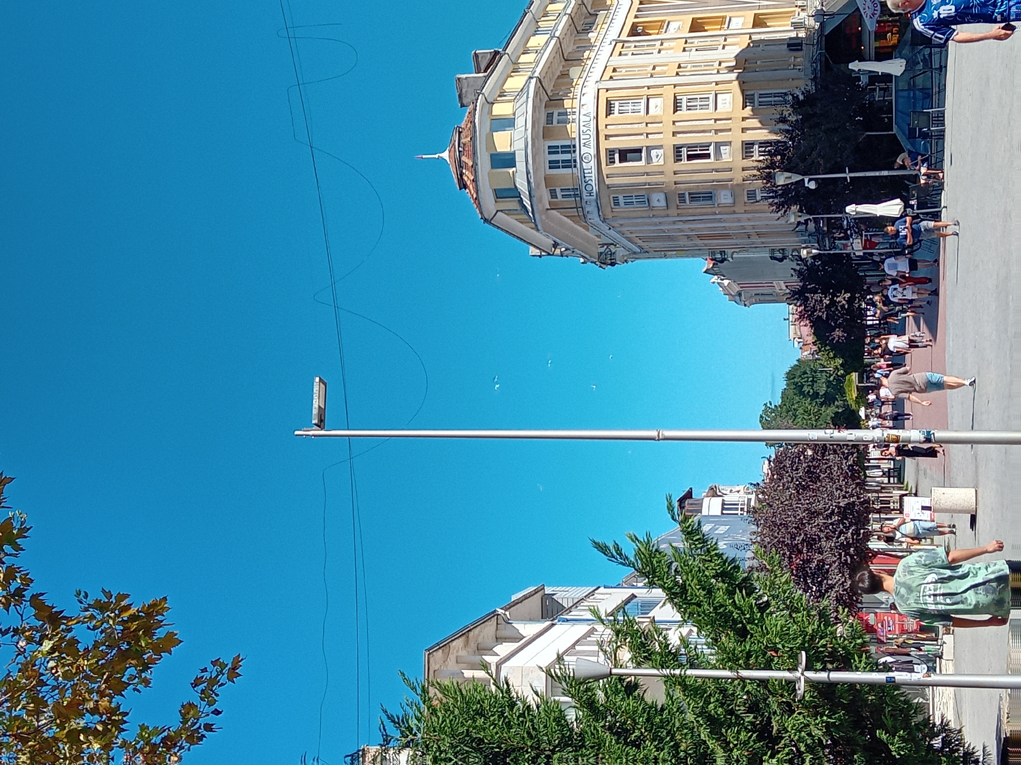 This image depicts a view of a pedestrian street in Varna, Bulgaria, likely on the pedestrianized section of Preslav Street, given the architectural style of the buildings and the general urban setting. The scene is characterized by a bright, clear blue sky with some faint white lines, possibly power lines or telephone wires, stretched across it. In the foreground, a tall, modern street lamp stands prominently in the center. To the left, a leafy green tree with dark branches occupies the side of the image, with a portion of a white building visible behind it.
Further down the street, on the right side, a distinctive yellow building with a rounded corner and an ornate dome-like roof dominates the view. The building has multiple windows and balconies, and a sign that reads "HOSTEL MUSALA" is clearly visible on its facade. Several other buildings line the street, displaying a mix of architectural styles, with some appearing older and more traditional.
The street itself is paved and filled with pedestrians walking in both directions. People are dressed in casual summer clothing, suggesting warm weather. Some individuals are walking alone, while others are in groups. Among the people, a young person in a green tie-dye t-shirt is facing away from the camera, walking down the street. Further along, other individuals are visible, including a person in a blue jersey with the number 10 on the back, who is partially in the frame on the far right.
Shops and cafes appear to be situated along the street, with awnings and outdoor seating visible in some areas. The overall impression is a vibrant urban street scene on a sunny day.