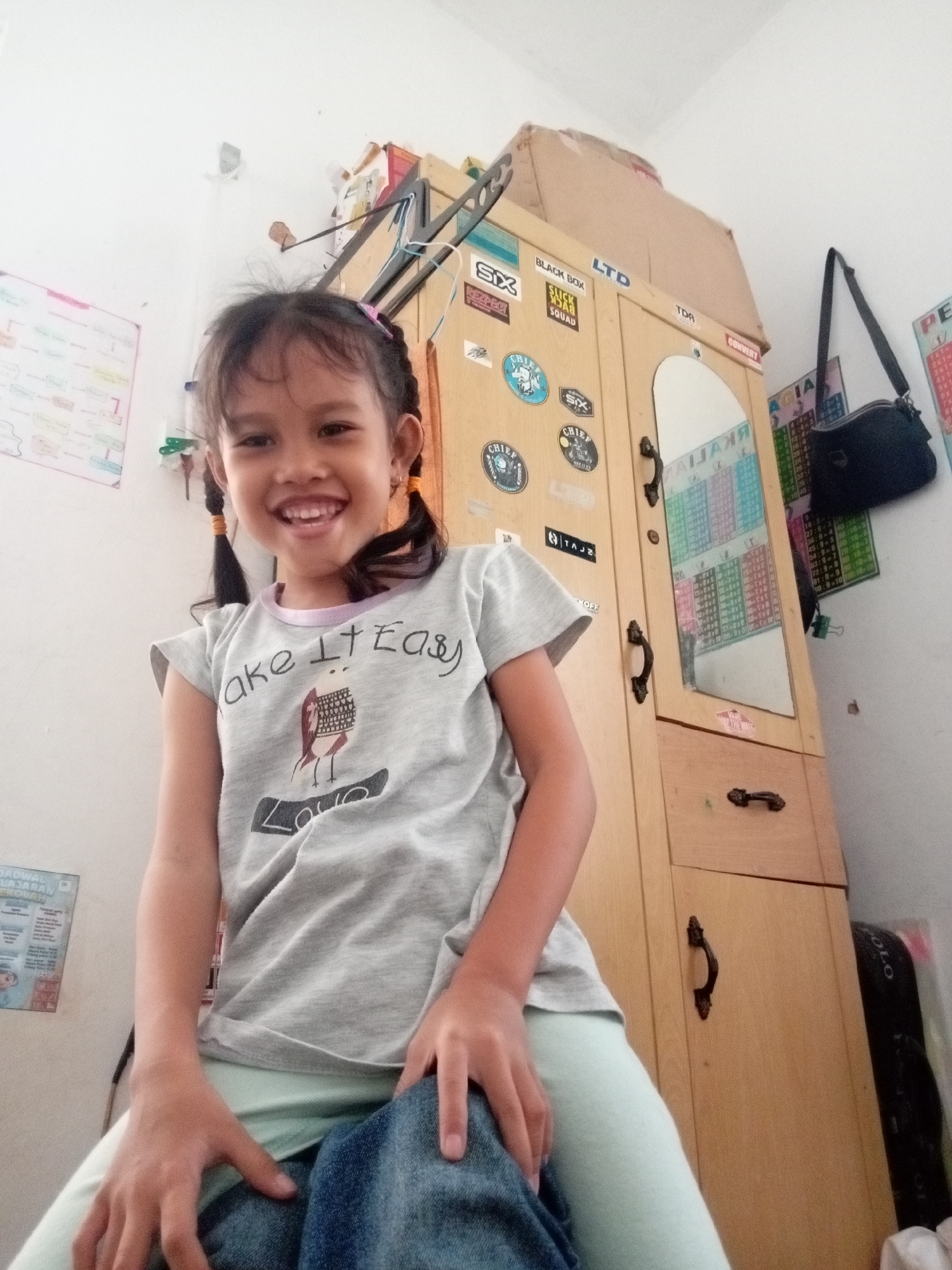 A young girl with pigtails, wearing a grey t-shirt with a graphic of a bird and text that reads "ake It Easy", is seated in front of a wooden cabinet. She is smiling broadly, with her teeth visible. Her hands are resting on her lap, which is covered by blue fabric, possibly jeans. The cabinet has two doors, one of which reflects the girl's image. The cabinet is adorned with various stickers, including those with "SIX", "CHIEF", "BLACK BOX", and "LTD". A black handbag hangs from the side of the cabinet. To the left of the girl is a whiteboard with what appears to be a mind map or diagram. The environment suggests an indoor setting, likely a bedroom or play area, in Batam, Indonesia.