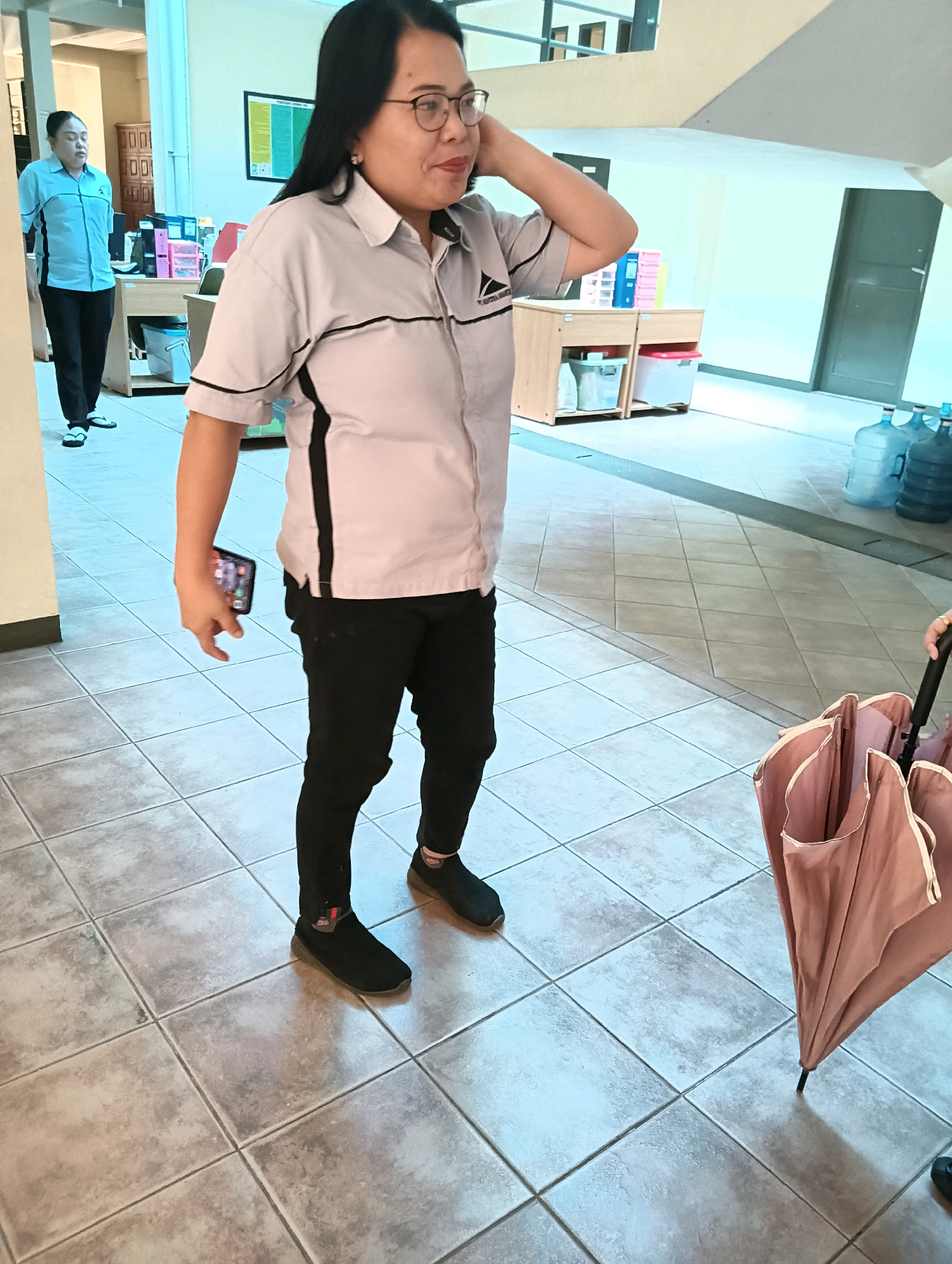 In an indoor hallway with tiled floors, a woman of Asian descent wearing glasses, a light grey short-sleeved collared shirt with dark trim, and black pants stands in the foreground, looking to her right and touching her head with her right hand. She is holding a dark-colored smartphone in her left hand, positioned in front of her. Beside her on the right, a closed pink umbrella with a black handle is visible. In the background, another woman in a light blue short-sleeved collared shirt and black pants walks away from the camera. The hallway contains several desks with office supplies, water cooler jugs, and an emergency exit sign. The scene appears to be within an office building or similar institutional setting in Tomohon, Indonesia.