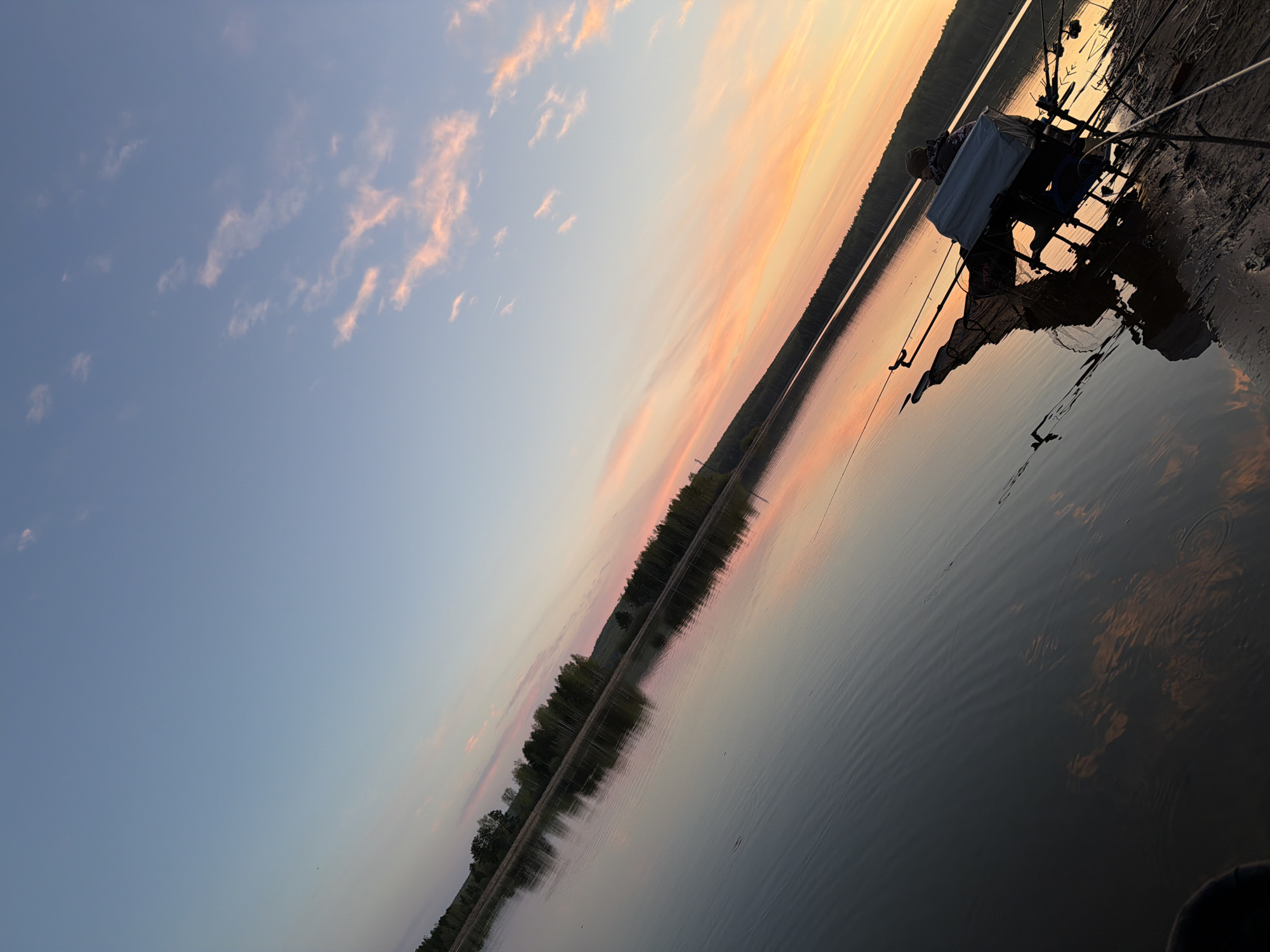 The image depicts a waterside scene, likely a river or lake. The photo is taken at a tilted angle. The sky above is a gradient of blue to light orange, with wisps of clouds in the blue section. A band of trees lines the shore opposite the viewer, reflected in the calm water. A person is fishing at the shoreline. Fishing rods and a large white container or cooler are near the person. The scene appears to be at sunset or sunrise due to the colors in the sky. It is not possible to confirm this image is from Chaykovsky, Russia, based on the image alone.