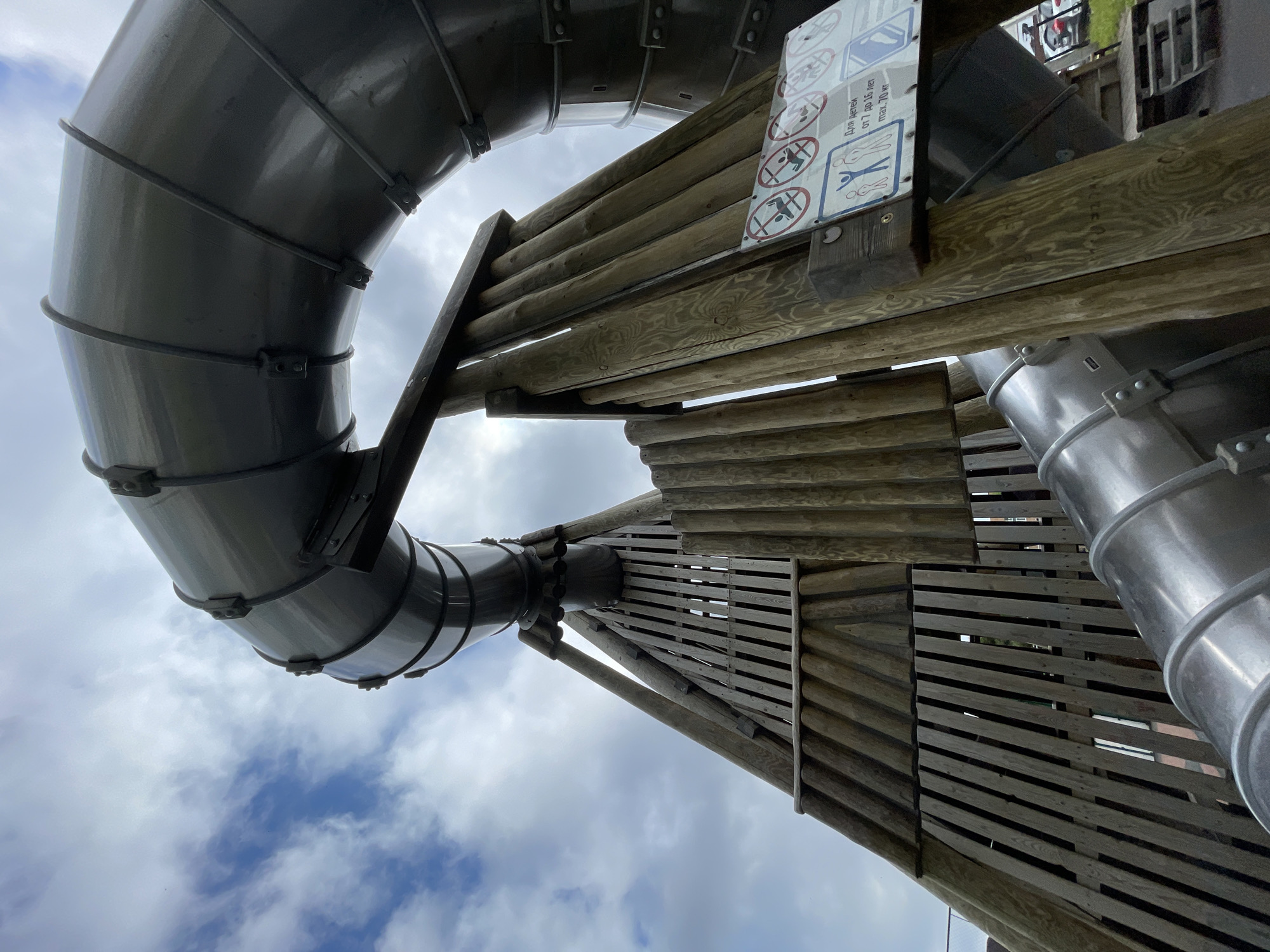 This low-angle photograph, taken in Izhevsk, Russia, captures a large, winding enclosed tube slide and its sturdy wooden support structure against a cloudy daytime sky. The scene depicts an outdoor recreational facility, likely a playground or adventure park.
The primary subjects are the slide and its wooden framework. The slide is constructed from segmented sections, alternating between dark gray/black and metallic silver, joined by visible bands and bolts. Its curved design suggests a twisting descent. The supporting structure is made of logs and planks of weathered wood, forming what appears to be a platform or tower from which the slide originates or is supported.
Attached to the wooden structure is a white informational sign with blue and red pictograms, indicating rules for the slide's use. Visible pictograms prohibit running, climbing on the outside of the slide, sliding head-first, and multiple people using it at once. Below these symbols, Russian text reads: "ДЛЯ ДЕТЕЙ от 7 до 16 ЛЕТ" (FOR CHILDREN from 7 to 16 YEARS OLD) and "max. 70 кг" (maximum 70 kg), clearly defining the age and weight restrictions for users.
The time of day is daytime, evidenced by the bright sky, which is filled with white and gray clouds, though patches of blue sky are visible. No people are present, so no specific activity is taking place, but the structure's purpose is evident. The image is devoid of any strong emotional cues, presenting a factual view of the playground equipment.