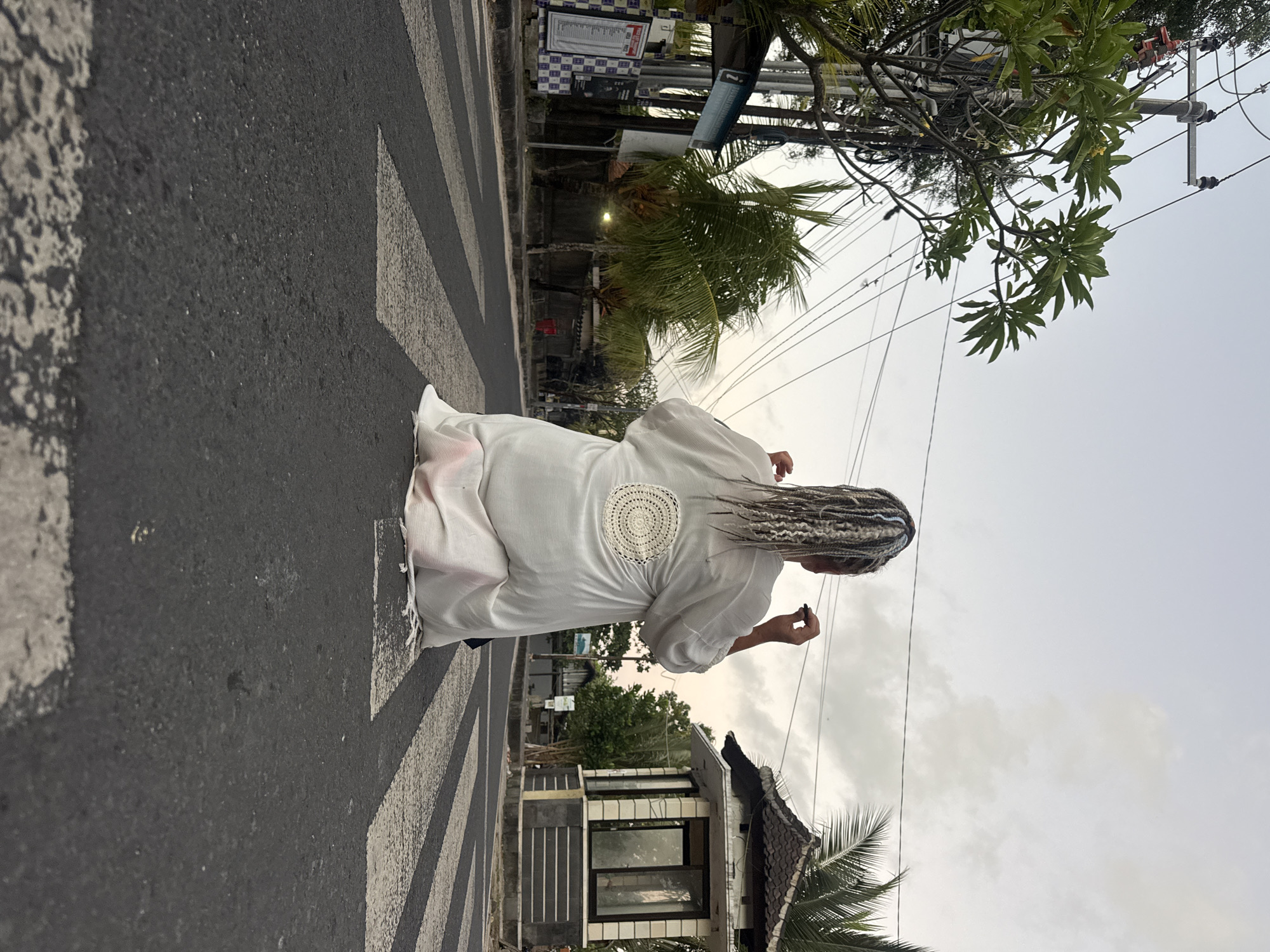 A person with blonde and gray braided hair is kneeling on a pedestrian crossing in Kuta, Indonesia. The individual is wearing a white, flowing robe with a circular crochet design on the back. The person is facing away from the camera, with their right hand raised, holding a dark object, possibly a pen or marker. In the background, there is a street with palm trees, power lines, and a small, traditional-style building with a dark roof. The sky is overcast, suggesting either early morning or late evening. The pedestrian crossing is marked with thick black and white stripes on asphalt.