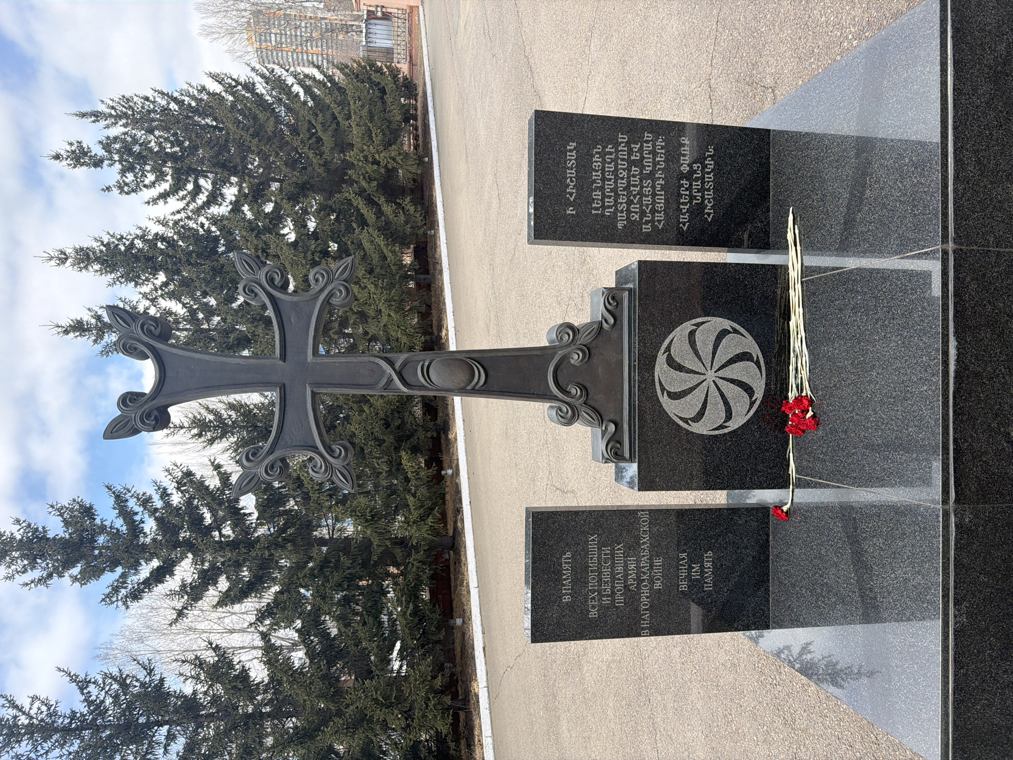 The image displays a solemn outdoor memorial located in Krasnoyarsk, Russia, dedicated to Armenians who perished or went missing during the Nagorno-Karabakh War.
Central to the memorial is a tall, dark, ornate metal Armenian cross (khachkar-style) with intricate swirling patterns on its arms and top, mounted on a polished black granite base. Flanking this central cross are two polished black granite steles. The stele on the left displays text in Russian, while the one on the right features text in Armenian, both conveying a message of remembrance.
At the base of the memorial, on a smaller granite slab carved with a traditional Armenian eternity symbol (Arevakhach), several red carnations and white slender flowers are reverently laid, serving as a poignant tribute. The immediate ground around the memorial is paved with light grey asphalt.
In the background, dense evergreen trees rise tall, with glimpses of a cloudy, light blue sky visible above them. Further in the distance, parts of a building can be discerned, suggesting an urban or semi-urban setting. The lighting indicates it is daytime, likely late morning or early afternoon, under a mixed sky.
The Russian inscription reads: "В ПАМЯТЬ ВСЕХ ПОГИБШИХ И БЕЗВЕСТИ ПРОПАВШИХ АРМЯН В НАГОРНО-КАРАБАХСКОЙ ВОЙНЕ ВЕЧНАЯ ИМ ПАМЯТЬ" (In memory of all the fallen and missing Armenians in the Nagorno-Karabakh War. Eternal their memory). The Armenian inscription reads: "Ի ՀԻՇԱՏԱԿ ԼԵՌՆԱՅԻՆ ՂԱՐԱԲԱՂԻ ՊԱՏԵՐԱԶՄՈՒՄ ԶՈՀՎԱԾ ԵՎ ԱՆՀԱՅՏ ԿՈՐԱԾ ՀԱՅՈՐԴԻՆԵՐԻ: ՀԱՎԵՐԺ ՓԱՌՔ ՆՐԱՆՑ ՀԻՇԱՏԱԿԻՆ:" (In memory of the fallen and missing Armenians in the Nagorno-Karabakh War: Eternal glory to their memory). The memorial stands as a profound symbol of remembrance and honor for the victims of the conflict.