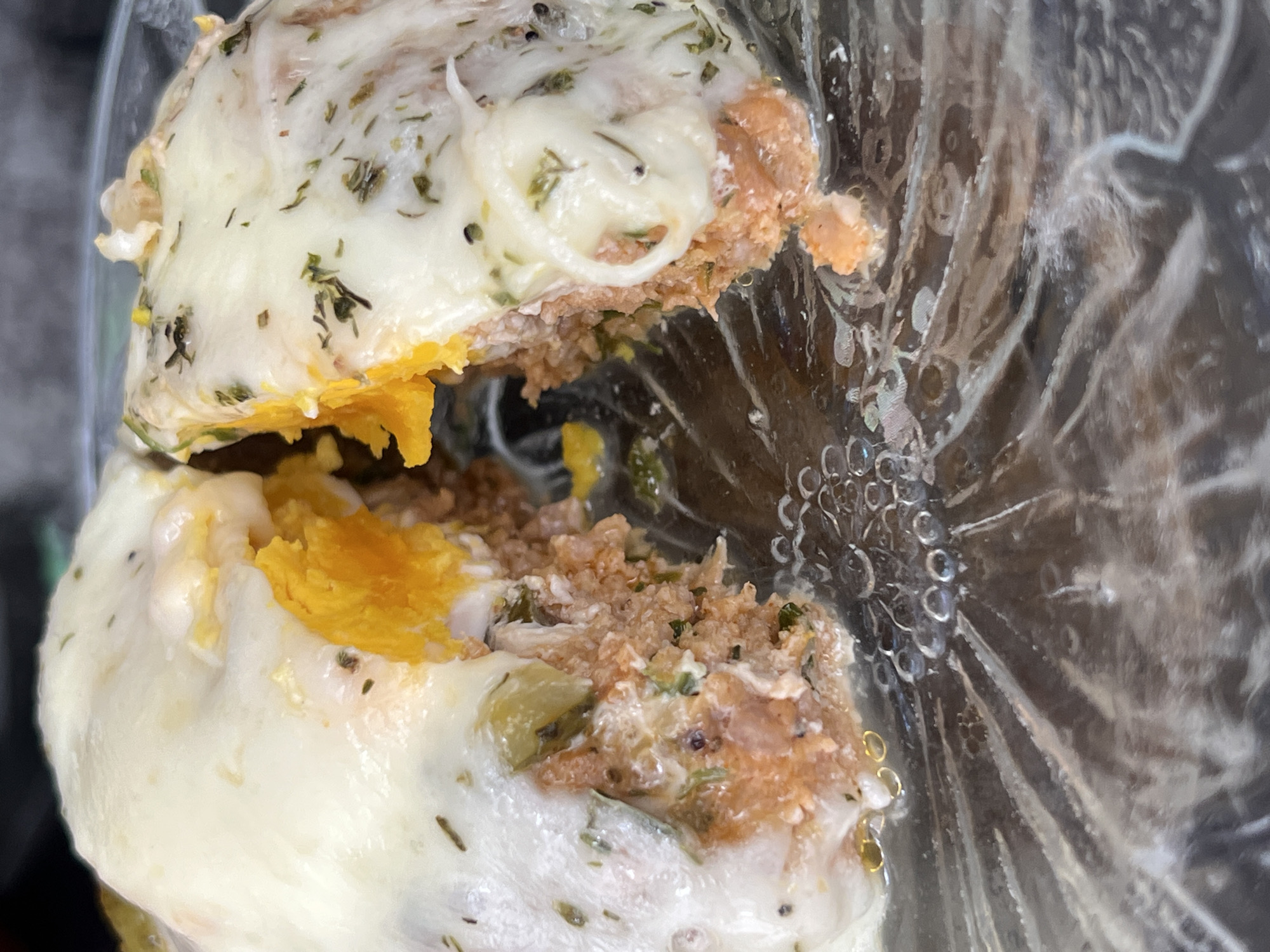 This image shows a close-up of a halved, filled food item, likely a stuffed chicken breast or similar dish. The filling appears to be a mixture of bread crumbs, herbs, and possibly other ingredients. A cooked egg yolk is visible within the filling. The food item is covered with melted cheese and herbs. The dish is contained within a plastic container. The location of this image is unknown.