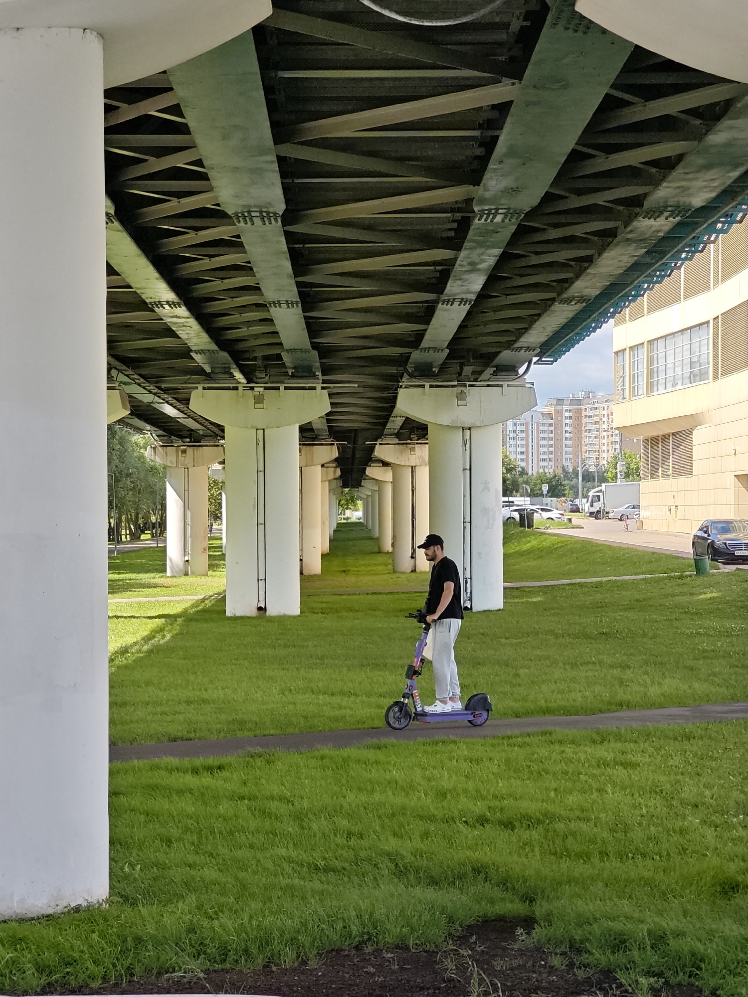 A person is riding an electric scooter on a paved path under a large concrete overpass in Moscow, Russia. The overpass is supported by numerous white concrete pillars. The ground beneath the overpass is covered with green grass, and a path winds through it. In the background, there is a building with a yellow facade and several apartment buildings. Several cars are parked in the background. The person on the scooter is wearing a black t-shirt, grey pants, and a black baseball cap. The scooter is purple and has white accents. The lighting suggests it is daytime, possibly late afternoon given the shadows.