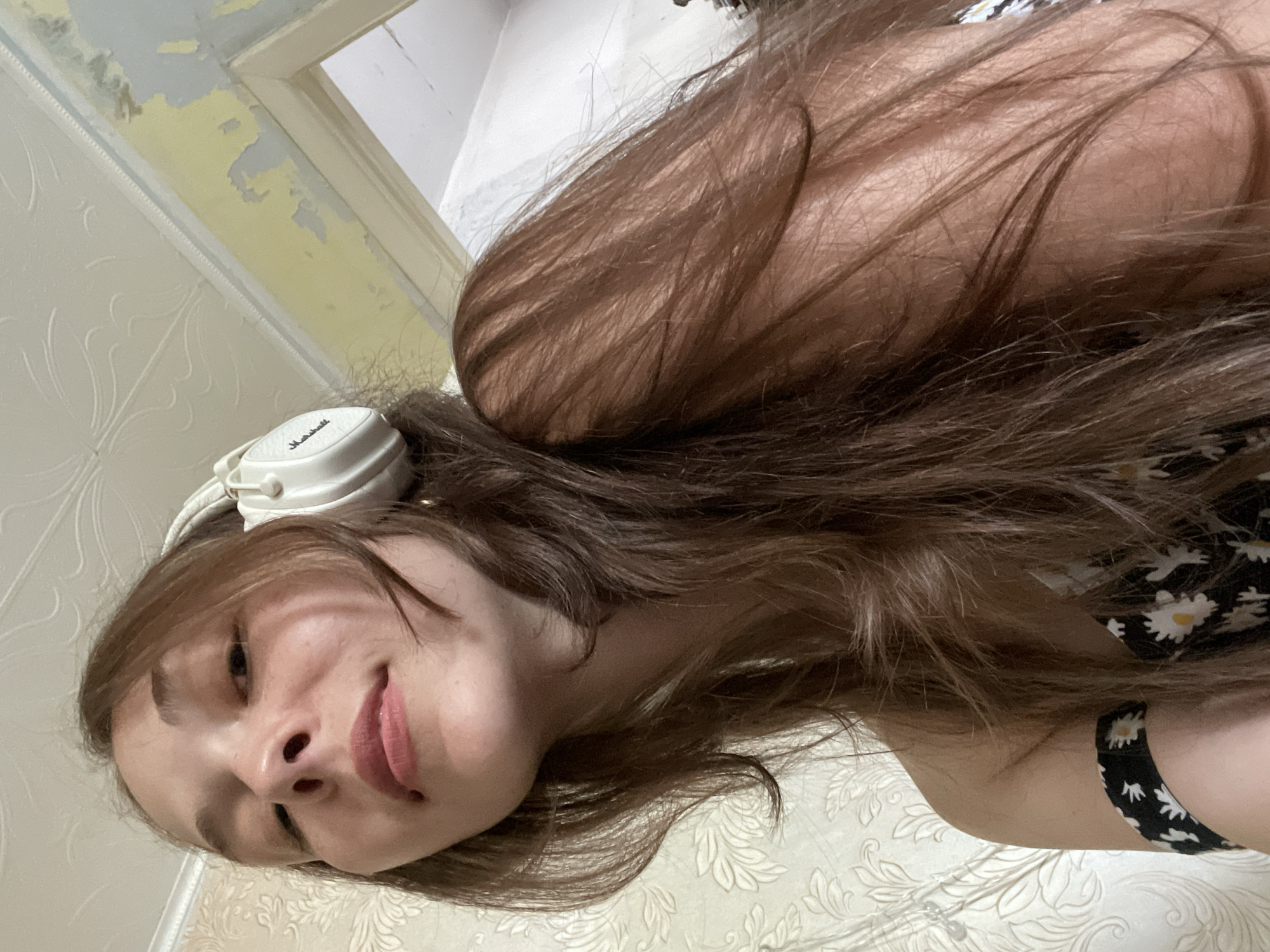 An overhead, close-up shot shows a young woman with long, brown hair wearing white headphones. She is looking down and to the left, with her mouth slightly open. She is wearing a black and white floral-patterned top. The background consists of a patterned wallpaper and a doorway with peeling yellow paint. The scene appears to be indoors.