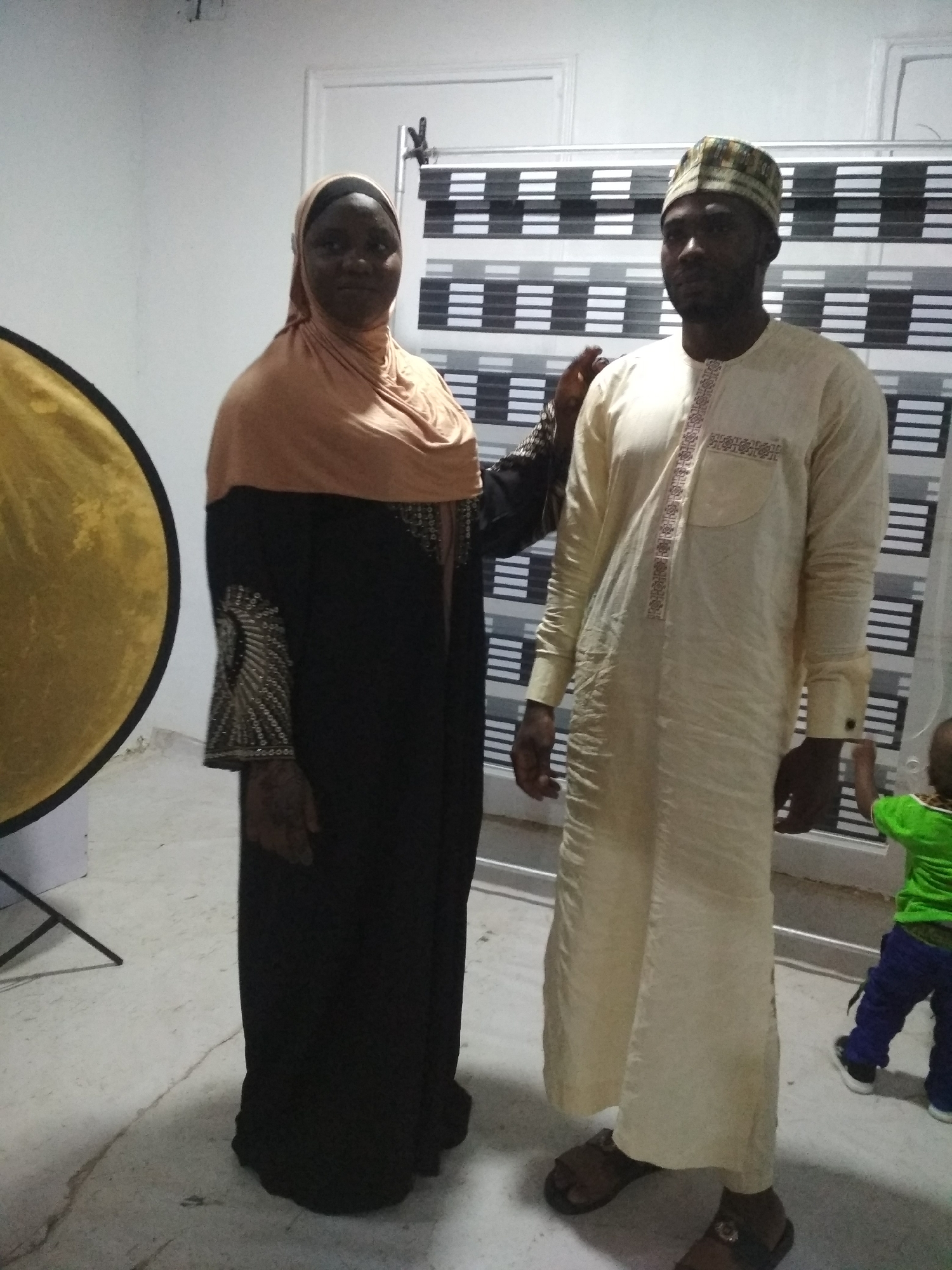 Two adults are standing in front of a black and white horizontally striped background. A woman on the left, wearing a black abaya with embellished sleeves and a tan hijab, has her left hand on the right shoulder of a man who is wearing a long off-white tunic and a patterned skullcap. A child is partially visible in the lower right corner, wearing a green shirt and blue pants. A large round gold reflector is to the left of the woman. The location is Unknown, Unknown.