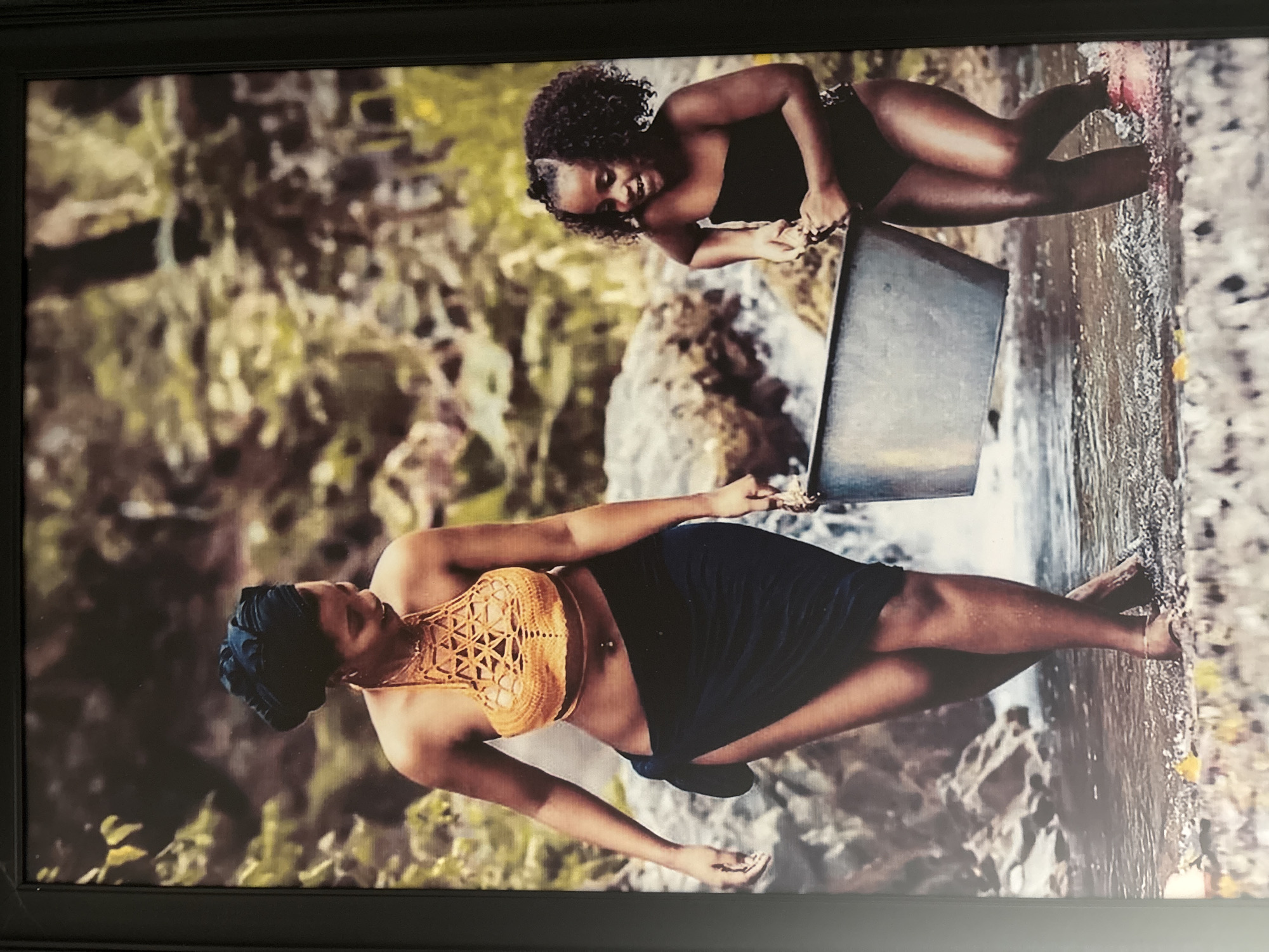 A framed photograph, oriented vertically, captures two women in a natural outdoor setting, likely a stream or river, surrounded by lush green foliage and rocks. The environment suggests a tropical or subtropical landscape, consistent with the Abuja, Nigeria context.
In the foreground, a woman stands in shallow water, facing slightly away from the camera and towards the right. She wears a striking golden-orange crocheted crop top with an intricate geometric pattern and a dark-colored wrap skirt. Her hair is covered by a dark headwrap, and a belly button piercing is visible. Her posture is relaxed, with her arms slightly bent.
Further back and to the right, a second woman is depicted crouching or kneeling in the water. She is looking directly at the camera with a broad, joyful smile. She has dark, curly hair and is dressed in a dark one-piece swimsuit or top. She holds a rectangular, dark-colored container or pan in the water, appearing to interact with it, possibly scooping or collecting water.
Both women are barefoot. The scene is brightly illuminated by natural daylight, conveying a serene and happy mood, primarily evidenced by the smiling woman. No visible text is present in the image.