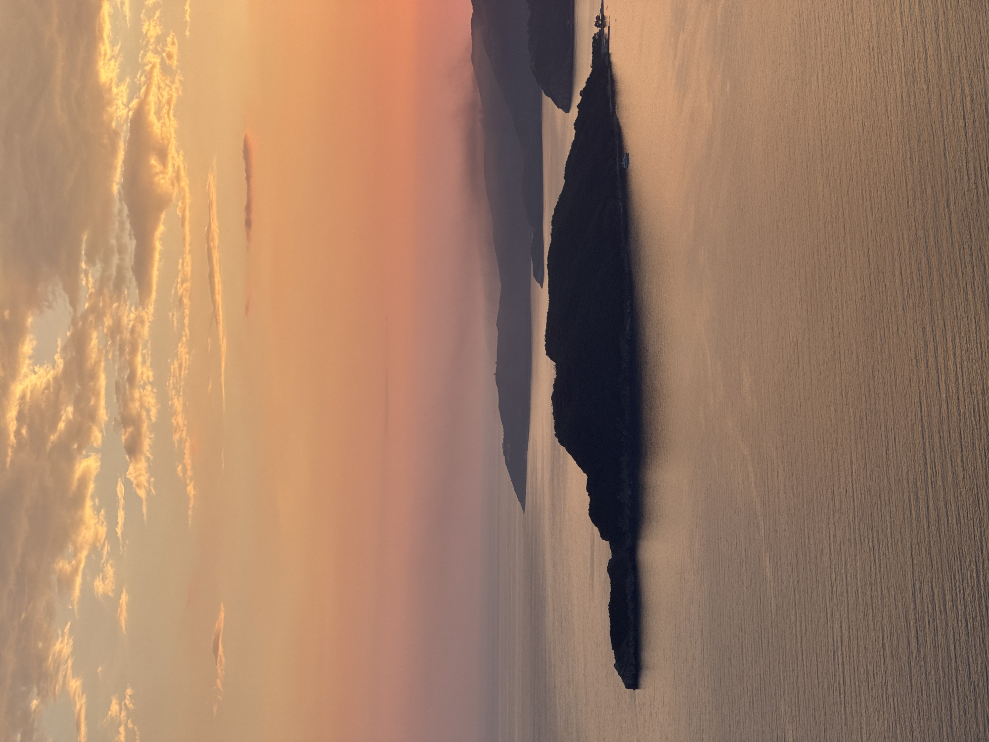 The image depicts a landscape at Sveti Stefan, Montenegro, showing the Adriatic Sea at dusk. In the foreground, an elongated, dark landmass with jagged edges juts out into the sea, possibly Sveti Stefan island or another coastal feature. The water displays a subtle rippling texture, illuminated by the soft, warm light. Beyond the island, outlines of hills and mountains can be seen in the distance, fading into the horizon. The sky transitions from a bright, hazy orange near the horizon to a lighter hue higher up, with scattered clouds adding texture.