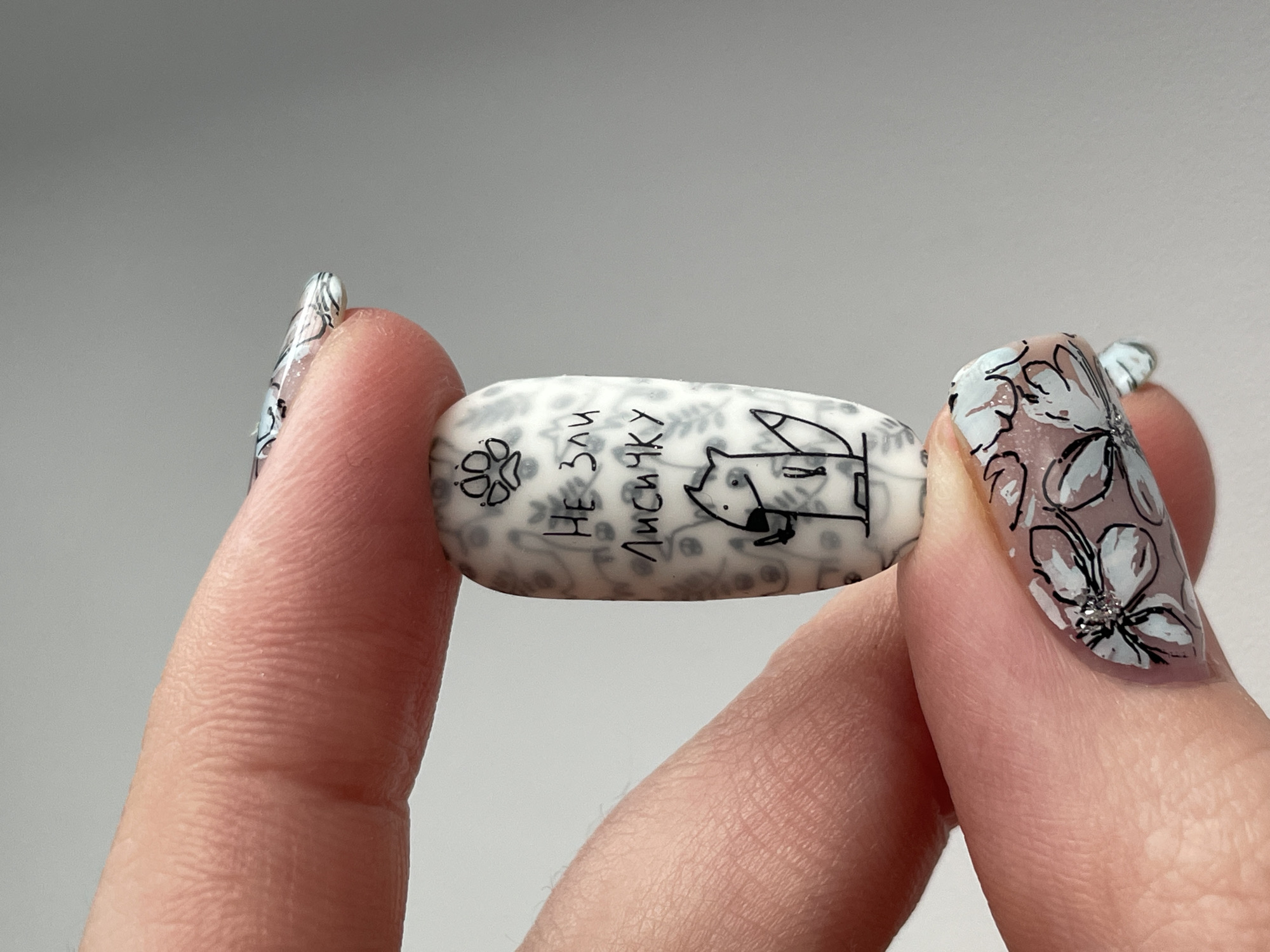 A close-up shot features a person's hands holding a small, white, elongated rectangular object with rounded edges. The object is held horizontally between the thumb and middle finger of the right hand and supported by the index finger of the left hand. The person's fingers are light-skinned, showing visible skin texture and creases.
Their fingernails are long, with an oval or almond shape, and are elaborately decorated. The nail art features hand-painted floral designs in black outlines, white, and pale blue/grey tones. Some nails are adorned with small silver glitter or rhinestone accents, particularly at the center of the flowers.
The white object itself has a subtle, light grey, intricate pattern underneath darker black line art. On the left side of the object, a paw print design is visible. Towards the center, Cyrillic text is written, with the words "НЕ ЗАЧ" (likely part of "НЕ ЗАЧЕМ" meaning "no need") and "ЛИСИЧКУ" (meaning "little fox," accusative case) clearly discernible. To the right, a stylized cartoon animal, resembling a fox or dog, is drawn in black outline, looking to the left with its tail visible.
The background is a plain, light grey, suggesting an indoor setting. The image captures the detailed nail art and the unique, custom-decorated object, possibly a piece of jewelry, a charm, or a decorative accessory. The scene takes place in Izhevsk, Russia.