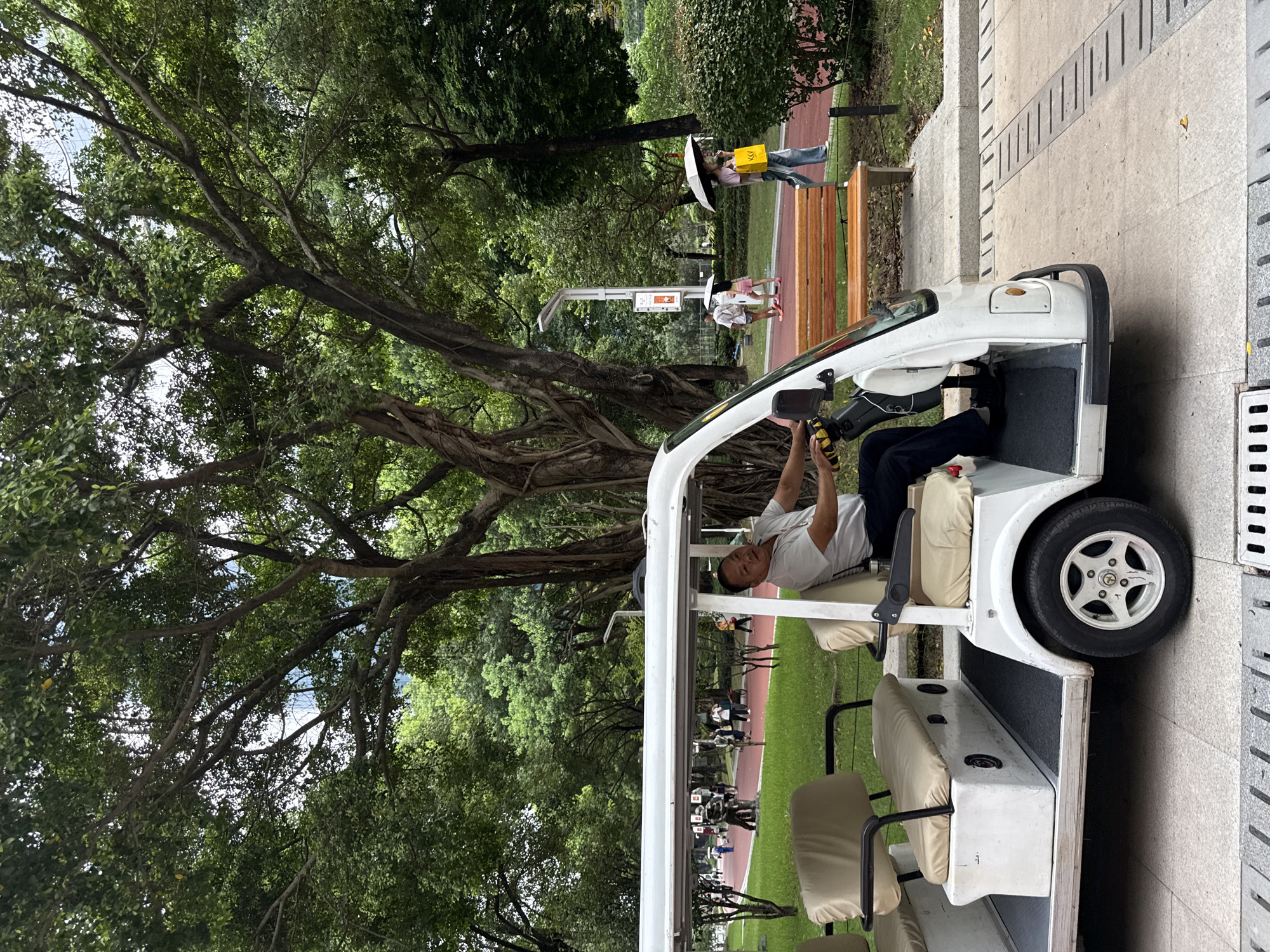A man drives a white electric golf cart through a park in Tianhe District, China. The man is wearing a light-colored polo shirt and dark pants, and is looking towards the camera with a slight smile. The golf cart has two rows of beige seats and a white roof. In the background, people are walking on a red pathway, and there are trees and bushes. One person is holding an umbrella. The ground is paved with light-colored stone tiles.