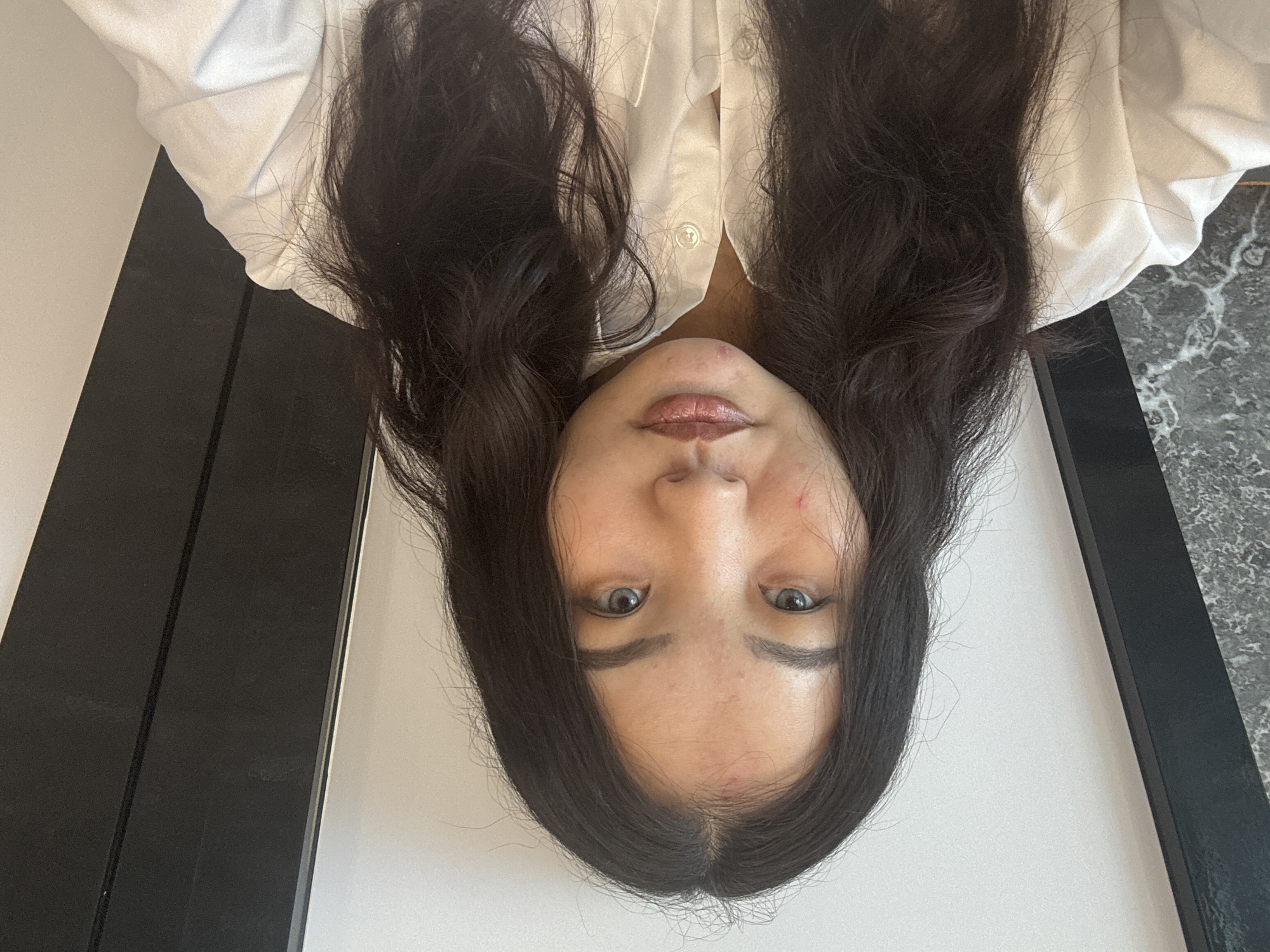 A medium shot, head-on photograph shows a young East Asian woman with dark, wavy hair and light blue eyes wearing a white button-down shirt. She is positioned in the center of the frame, facing directly towards the camera. The background consists of a light-colored wall on the right, with a dark, vertical element on the left side and a section of a patterned, dark surface. The lighting appears to be natural, coming from the front, casting soft shadows. There is a minor blemish on her forehead and a small red mark on her left cheek. Her lips are glossy and slightly parted. The overall scene suggests an indoor setting, possibly a home or studio. No specific location within Astana, Kazakhstan, can be determined from the visual information provided.