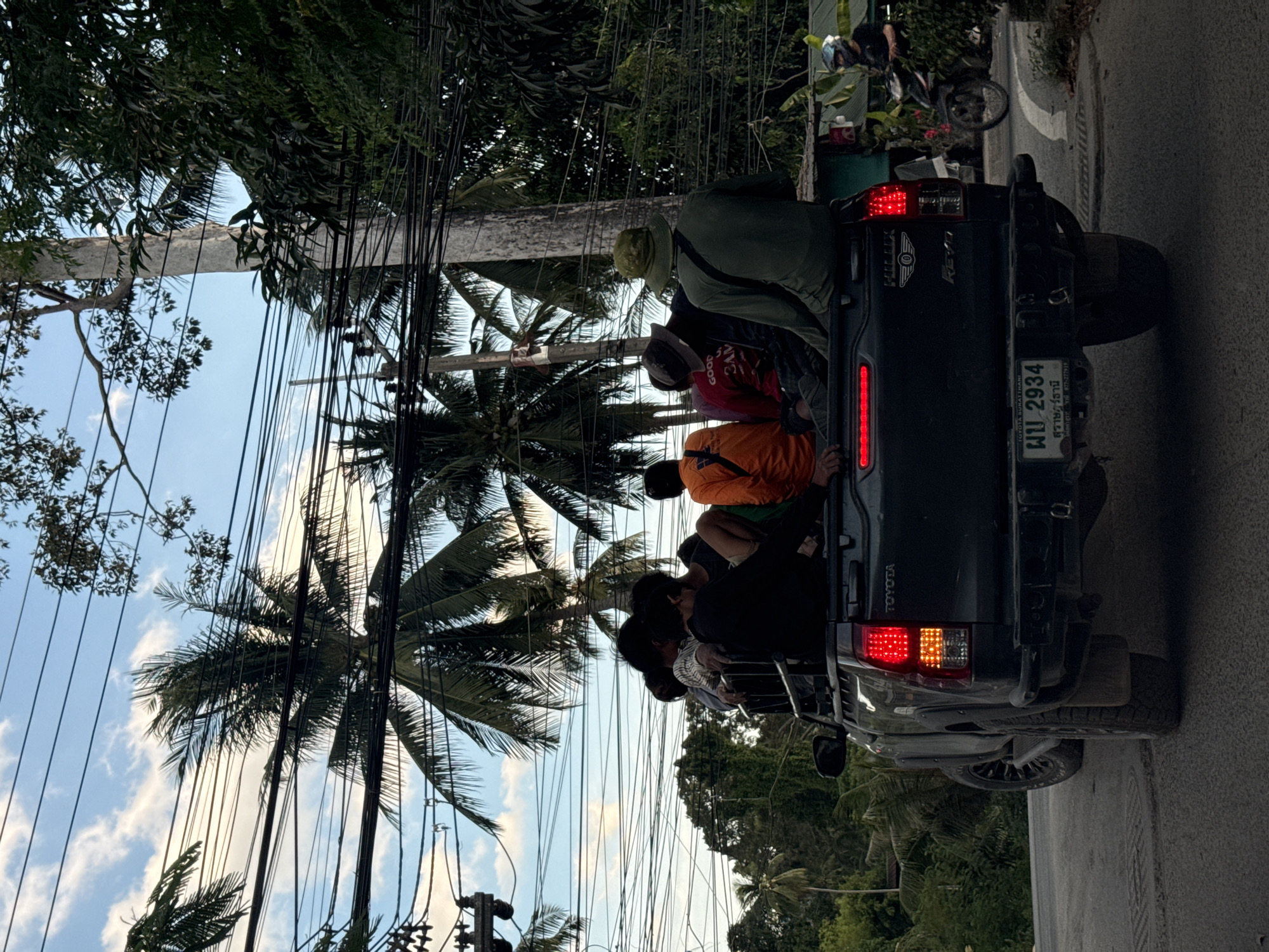 In Baan Mae Nam, Thailand, a dark-colored Toyota Hilux pickup truck is seen from behind, traveling on a paved road. The truck bed is occupied by several individuals. In the foreground, the rear of the truck is visible, with its taillights illuminated and the license plate reading "พบ 2934". A man wearing a green bucket hat and a green long-sleeved shirt is seated in the back, facing away from the camera. To his left, another person is seated, wearing a red shirt with white lettering that appears to read "GOOD SAM". An individual in an orange shirt is also visible. The background features a clear blue sky with scattered clouds, interspersed with numerous power lines. Tall palm trees and dense green foliage are present, indicating a tropical environment. To the right of the truck, a person on a motorcycle is seen passing by, wearing a helmet.