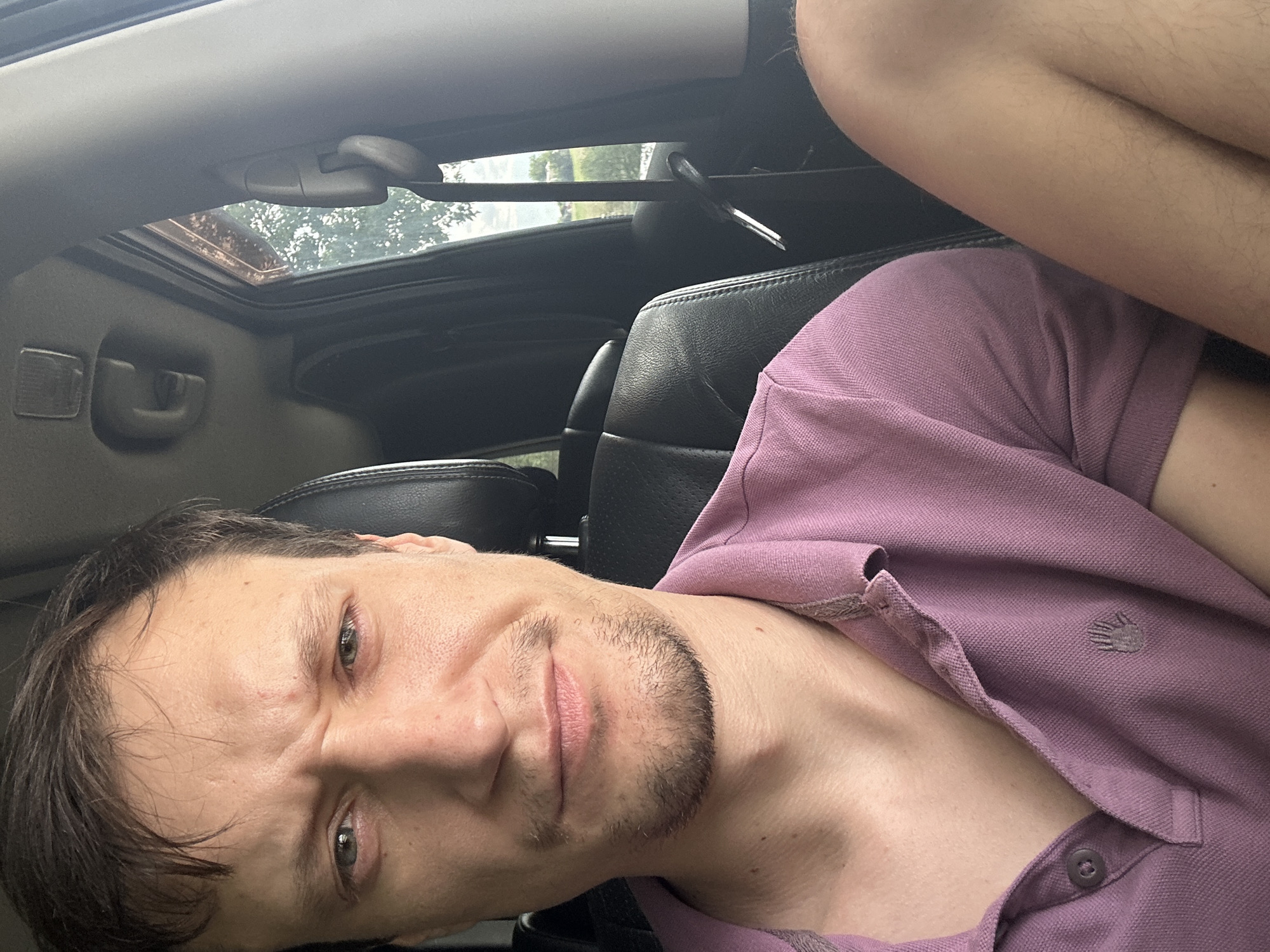 A man with light brown hair and a light beard is sitting in the driver's seat of a car. He is wearing a mauve polo shirt. His left arm is bent, and his hand is resting on his leg. The interior of the car appears to be dark leather. Outside the car window, trees and foliage are visible. The scene is likely in Makiv, Ukraine, given the context.