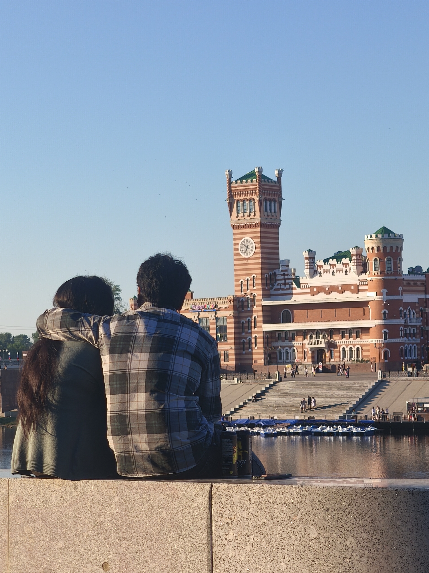 In Yoshkar-Ola, Russia, on a clear day, two individuals, a man and a woman, are seated from behind, embracing each other. The man, wearing a plaid shirt, has his arm around the woman, who has long dark hair and is wearing a green sweater. They are seated on a concrete ledge overlooking a body of water. In the background, a large, ornate red brick building with a clock tower and green-capped turrets stands prominently. The building is reminiscent of a castle or a historic European structure. The water in front of the building is dotted with several small boats. The overall scene suggests a peaceful and romantic moment in a scenic urban setting.
