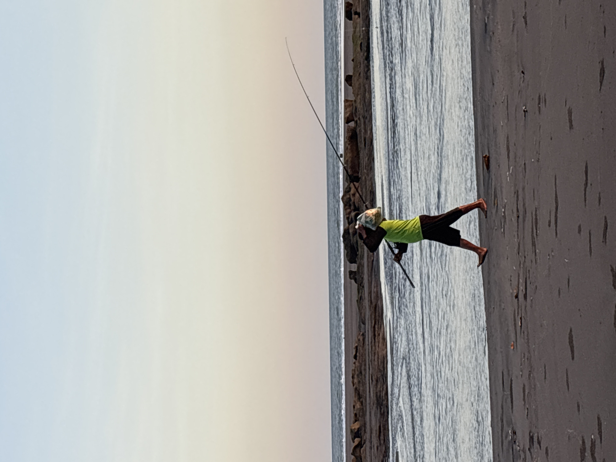 A man is fishing on a beach in Kerambitan, Indonesia. He is wearing a light green shirt and dark shorts and is standing on the dark sand near the water. He holds a fishing rod that is angled upwards towards the sky. The sea is visible to his left, and the sky above is a pale color, suggesting either dawn or dusk. A barrier of rocks separates the sand from the water.