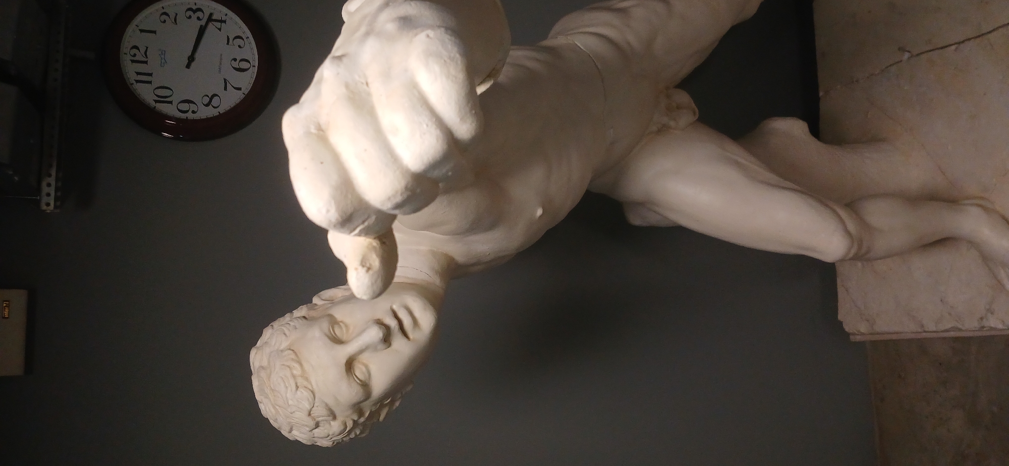 The image captures an indoor scene featuring a white, classical-style statue of a nude male figure. The statue, likely made of marble or plaster, is depicted in a dynamic pose, lunging forward with its right arm extended and hand clenched into a fist, pointing downwards. The figure's head is tilted, and its gaze is directed towards the extended fist. Intricate details of musculature and facial features are visible. The statue rests on a light-colored, rectangular pedestal, possibly made of stone, which shows some visible texture and cracks.
In the background, a plain dark grey wall provides a minimalist setting. A round wall clock with a dark wooden frame and a white face is hung in the upper left portion of the image. The clock's hands indicate the time is approximately 2:19. The word "QUARTZ" and the brand name "RHYTHM" are visible on the clock face, along with standard numerical hour markers. On the lower left section of the wall, a small, light-colored rectangular object displays the partial text "infor.T". A metallic, slotted rack or shelf is also faintly visible on the far left. The scene is illuminated by an unseen light source from the upper right, highlighting the statue and casting subtle shadows. This setting suggests a display within a museum, gallery, or private collection, potentially located in Moscow, Russia.