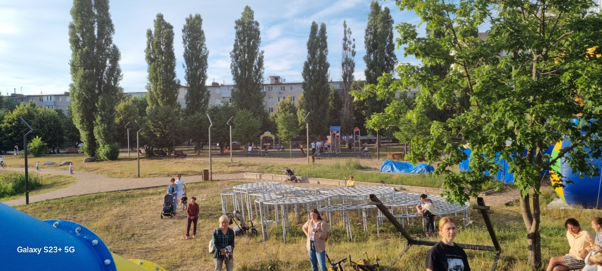 An outdoor scene captures a children's playground area in Shostka, Ukraine, on a clear day. Tall trees with dense green foliage frame the view, with a multi-story residential building visible in the background. Several people are scattered throughout the playground, some appearing to be adults supervising children, while others are engaged in their own activities.
In the foreground, a portion of a large, colorful inflatable structure, possibly a bouncy castle, is visible on the right side, predominantly blue with yellow and green accents. To the left, a section of a blue and yellow playground slide is also present. A paved pathway winds through the grassy area, with a few children on scooters seen in the distance.
Several metal structures, perhaps parts of new playground equipment or a decorative installation, are present in the mid-ground. A few strollers are parked or in use. Some people are standing, while others are seated. The ground is covered in dry grass and dirt. The sky is partly cloudy. The overall impression is a public park or recreational area in an urban setting.