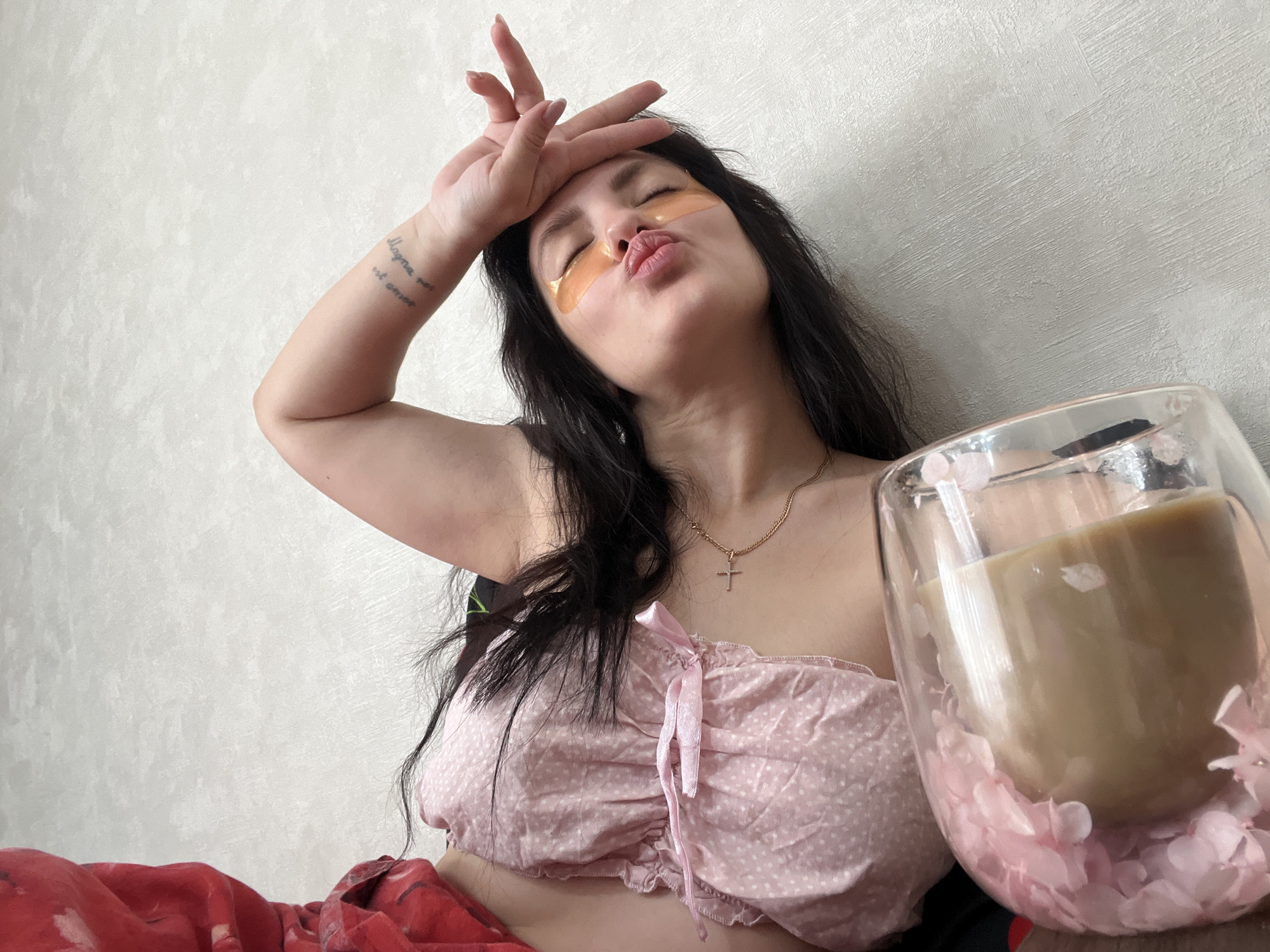 A young woman with long dark hair is captured in a close-up, indoor setting against a light-textured wall. She is engaged in a moment of self-care and playful expression, wearing hydrogel eye patches under her eyes. Her left hand rests casually on her forehead, fingers slightly splayed, while her eyes are closed, and her lips are pursed in a "kiss face."
She is dressed in a light pink, polka-dotted off-the-shoulder top with a ribbon tie. A thin gold chain necklace with a small cross pendant adorns her neck. On her left inner forearm, a tattoo is visible, reading "Magna pax est amor" (Latin for "Great peace is love"). Part of a red patterned blanket or fabric is seen on her lower body, suggesting she is seated and relaxing.
To her right, a clear, double-walled glass mug contains a light brown beverage, possibly coffee or a latte. The outer wall of the mug is decoratively filled with small pink, translucent elements resembling petals. The overall scene conveys a relaxed, casual, and personal atmosphere. The image is set in Марковский, Russia.
