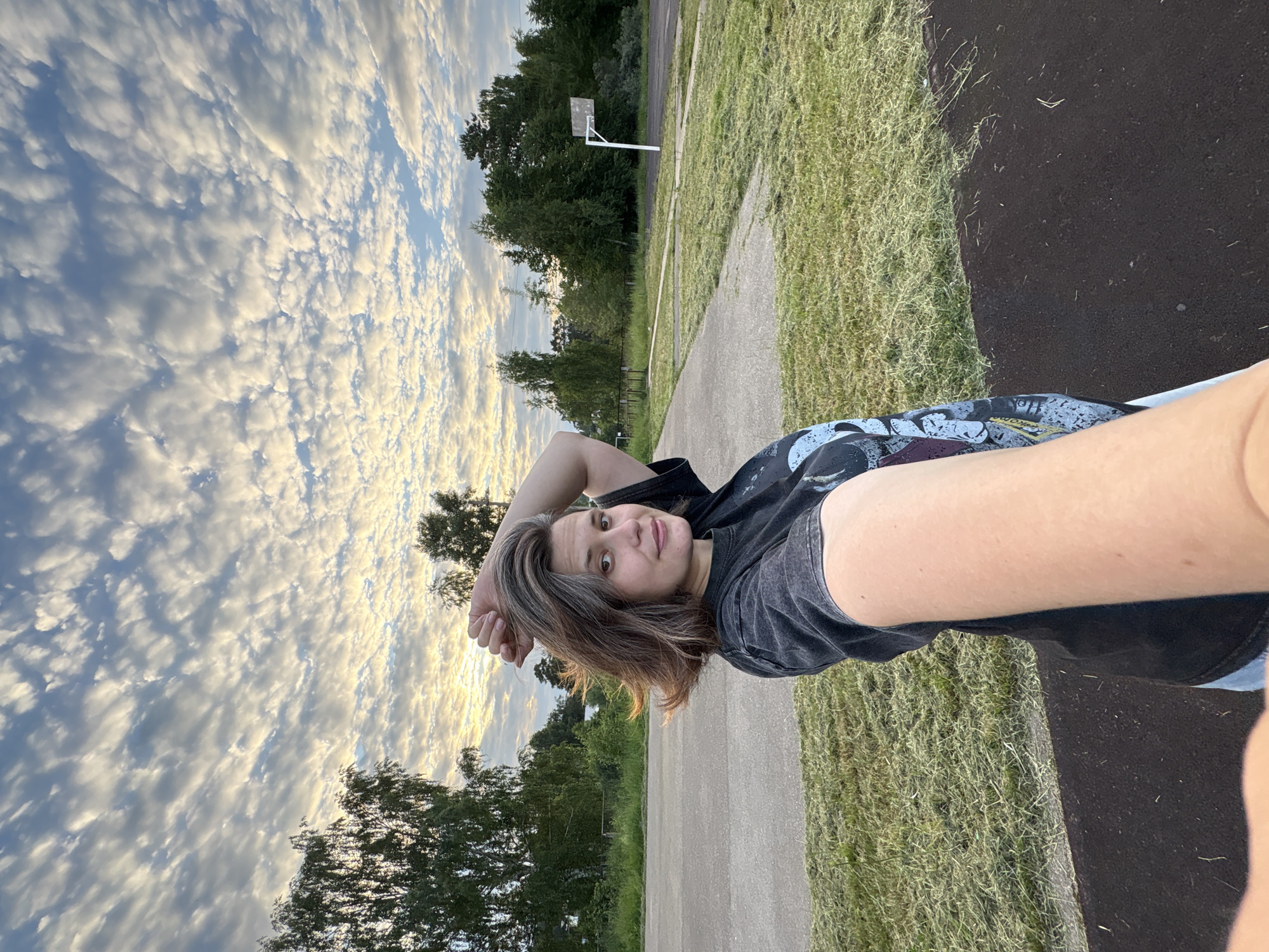 A woman is taking a selfie outdoors at dusk. She is standing in front of a basketball court with a hoop and a partially paved area. The sky is filled with a dramatic display of altocumulus clouds, illuminated by the setting sun. Trees and grass surround the court. The woman has brown hair and is wearing a dark t-shirt with a graphic print. Her arm is raised, holding the camera for the selfie. The background suggests an outdoor recreational area, possibly in a park or a schoolyard, within the city of Chaikovsky, Russia.