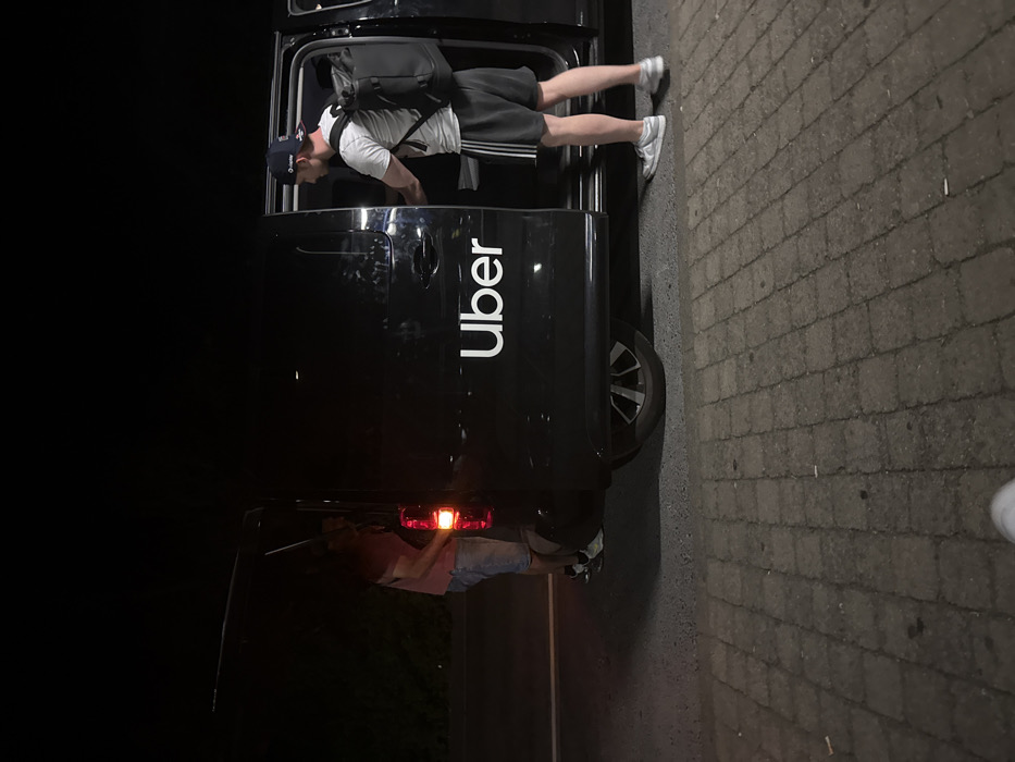 A man is reaching into the back of a black Uber vehicle. He is wearing a white shirt, dark shorts, a baseball cap, and a backpack. The vehicle is parked on a brick surface, and the passenger side door is open. The rear door is also ajar, and another person is possibly visible in the back. The scene is poorly lit, with the rear light illuminated.