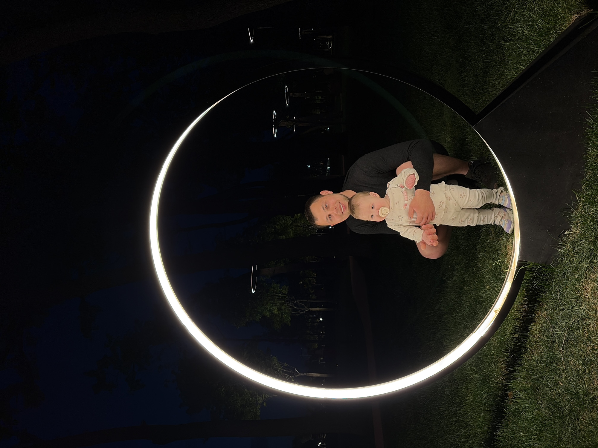 A medium shot depicts a man holding a baby inside a large, illuminated ring structure in what appears to be an outdoor location. The man, fair-skinned with dark hair, wears a dark-colored shirt. He is cradling the baby, who is dressed in light-colored pajamas, possibly with a floral pattern.
The light emitted from the circular structure illuminates the man and baby. The grass and ground underneath the ring are visible. In the background, vague outlines of trees and other elements are discernible, suggesting the scene is at nighttime.
There is no verifiable indication that the photo was taken in Krasnodar, Russia.