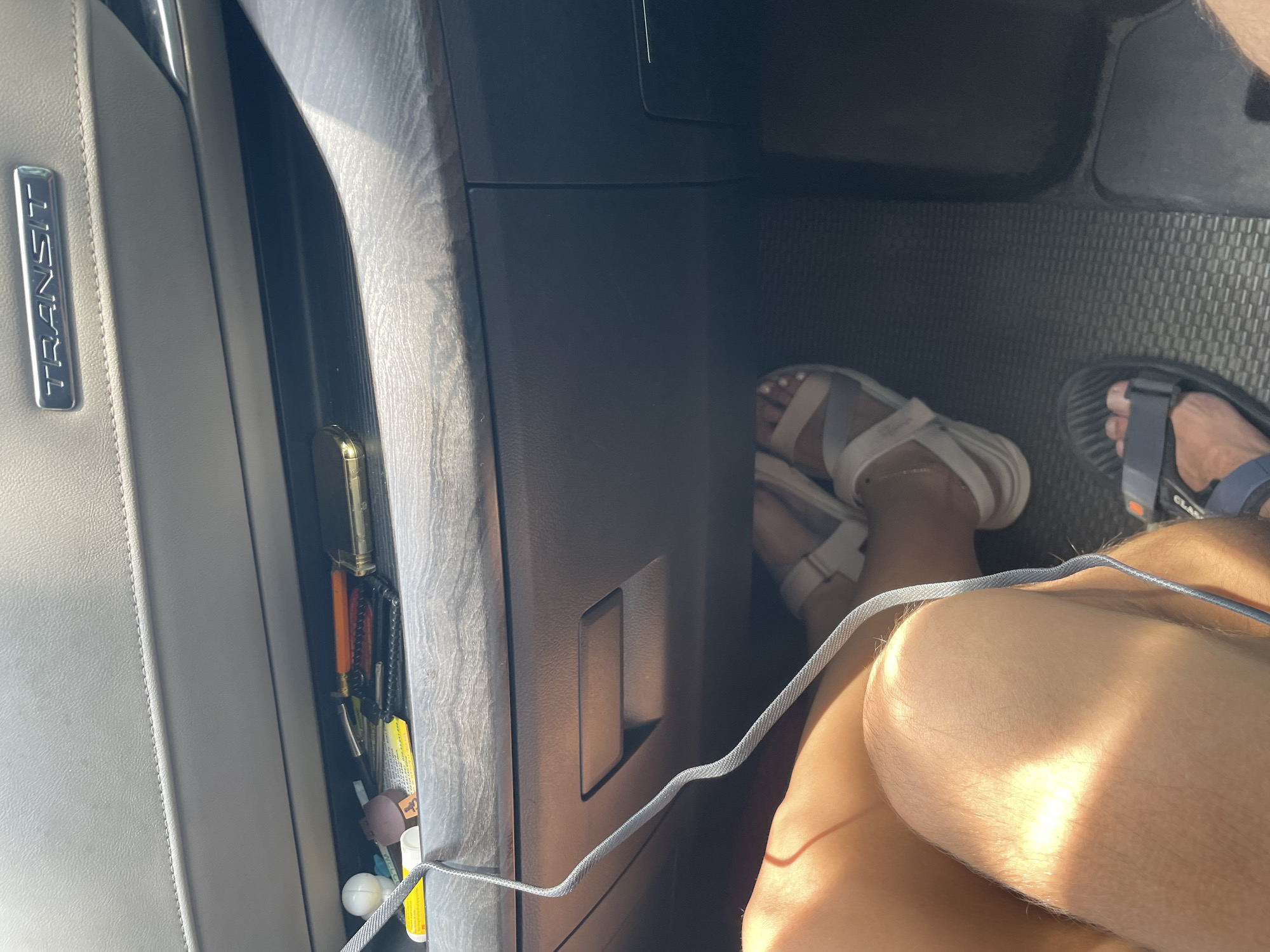 An overhead shot shows a person's legs and feet inside a vehicle. The vehicle appears to be a Ford Transit van, as indicated by the "TRANSIT" emblem on the dashboard. The person is wearing sandals and has their legs crossed. A charging cable snakes down from the dashboard, where a collection of items, including keys and what appears to be a small notebook, are stored in a compartment. The lighting suggests it is daytime, and the interior of the vehicle is clean and organized. The context suggests this scene takes place within the city of Thủ Đức, Vietnam, likely on a journey or during a stop.