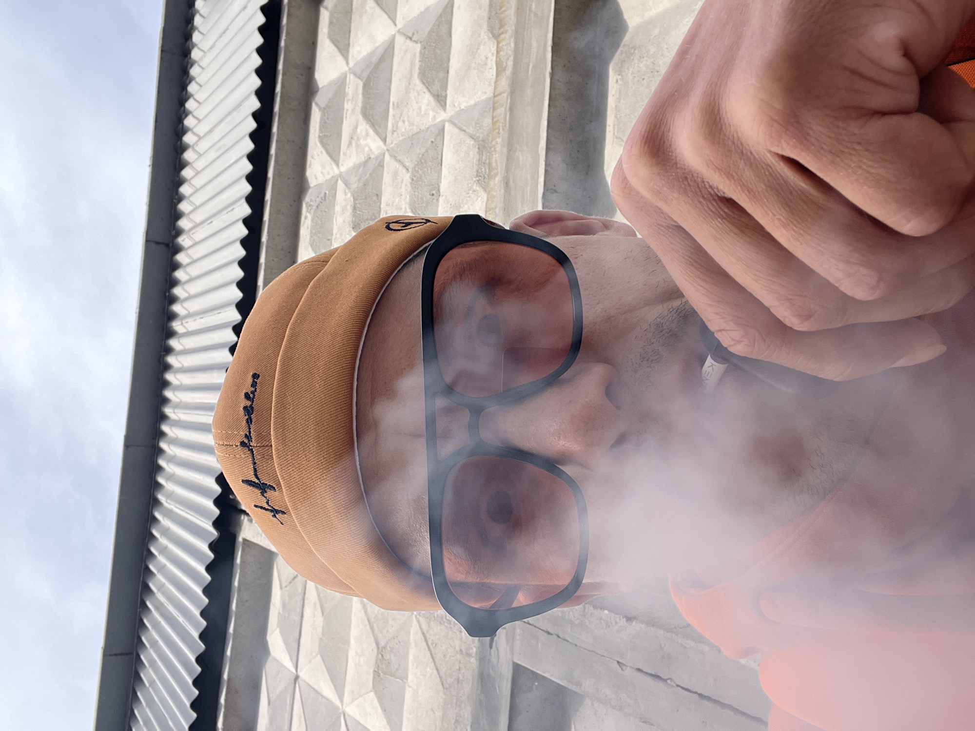 A person is captured in a low-angle shot, exhaling a dense cloud of vapor. The subject, likely male, is wearing a light brown or tan beanie with black stylized embroidery or text, black-framed sunglasses with orange-tinted lenses, and an orange or rust-colored hooded garment. Visible facial features include stubble on the chin and jawline. One hand is partially visible on the right side of the frame, clenched and holding a white vaping device that has the word "KEY" printed on it. The vapor originates from the person's mouth and obscures the lower part of their face.
The background on the left features a clear blue sky with minimal cloud cover, next to a vertical corrugated metal structure. On the right, behind the person, is a textured concrete wall with a repeating geometric pattern, suggesting a modern or industrial architectural element. The lighting is bright, indicating it is daytime, with clear shadows. The image captures a moment of vaping activity outdoors in Chaikovsky, Russia.