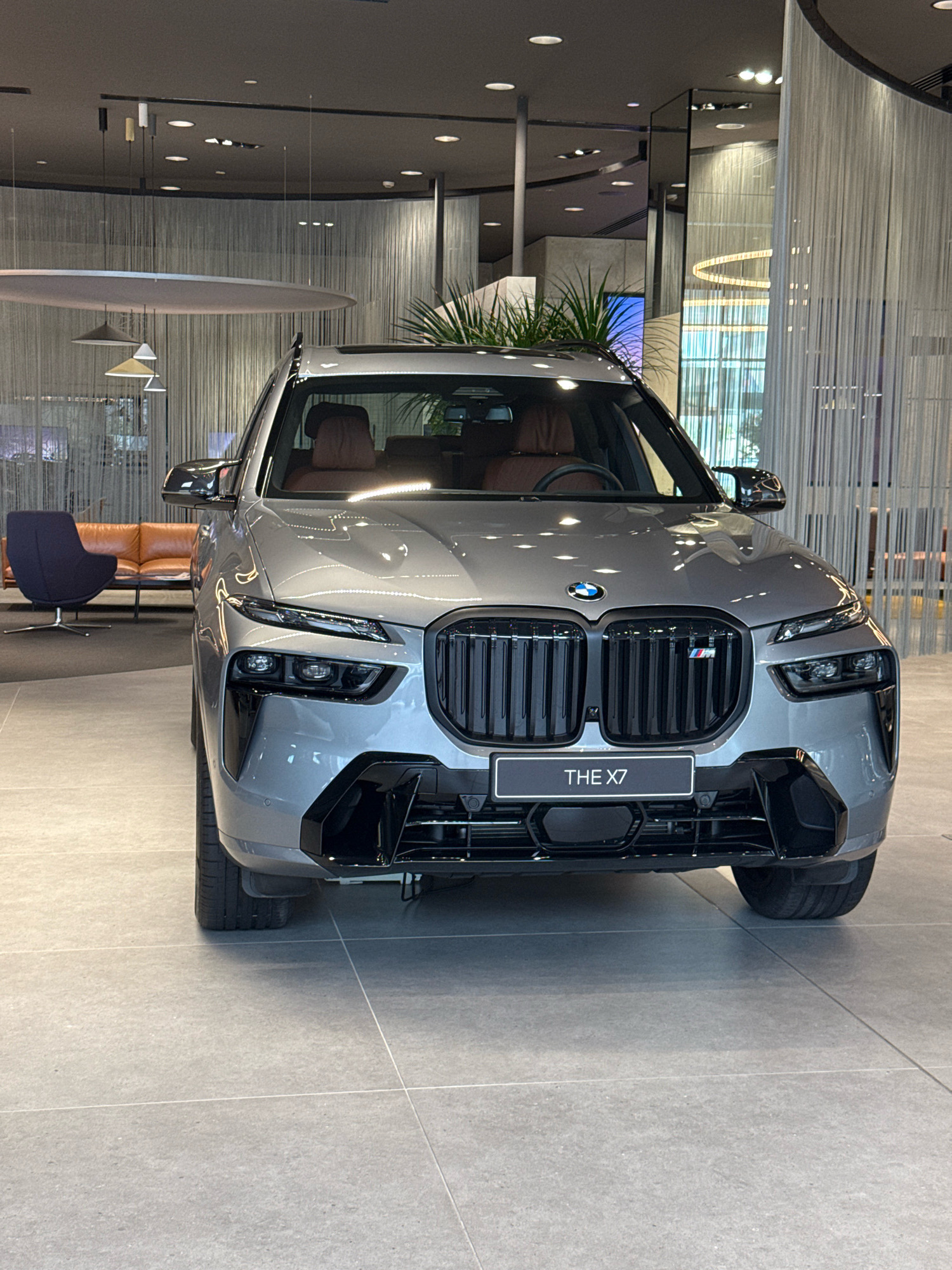 The image displays a front-facing view of a grey BMW X7 car parked indoors on a tiled floor. The car has a large black grille with the BMW logo at the top center. A license plate with "THE X7" is visible below the grille. The vehicle has sharp, modern headlights and a sporty front bumper. The interior of the car, with brown leather seats, is partially visible through the windshield.
The environment appears to be a modern showroom or dealership, suggested by the sleek ceiling with recessed lighting and decorative hanging lights. In the background, a large, curved wall covered with vertical white fabric or streamers adds to the sophisticated ambiance. To the left, a minimalist blue armchair with a swivel base and a low, rectangular coffee table are arranged near a tan leather sofa. A potted plant with green foliage is also visible in the background, adding a touch of nature to the space. The overall lighting is subdued and ambient, typical of a high-end retail or exhibition space. There are no people present in the image. The scene does not contain any specific markers to place it in Dubai, United Arab Emirates.