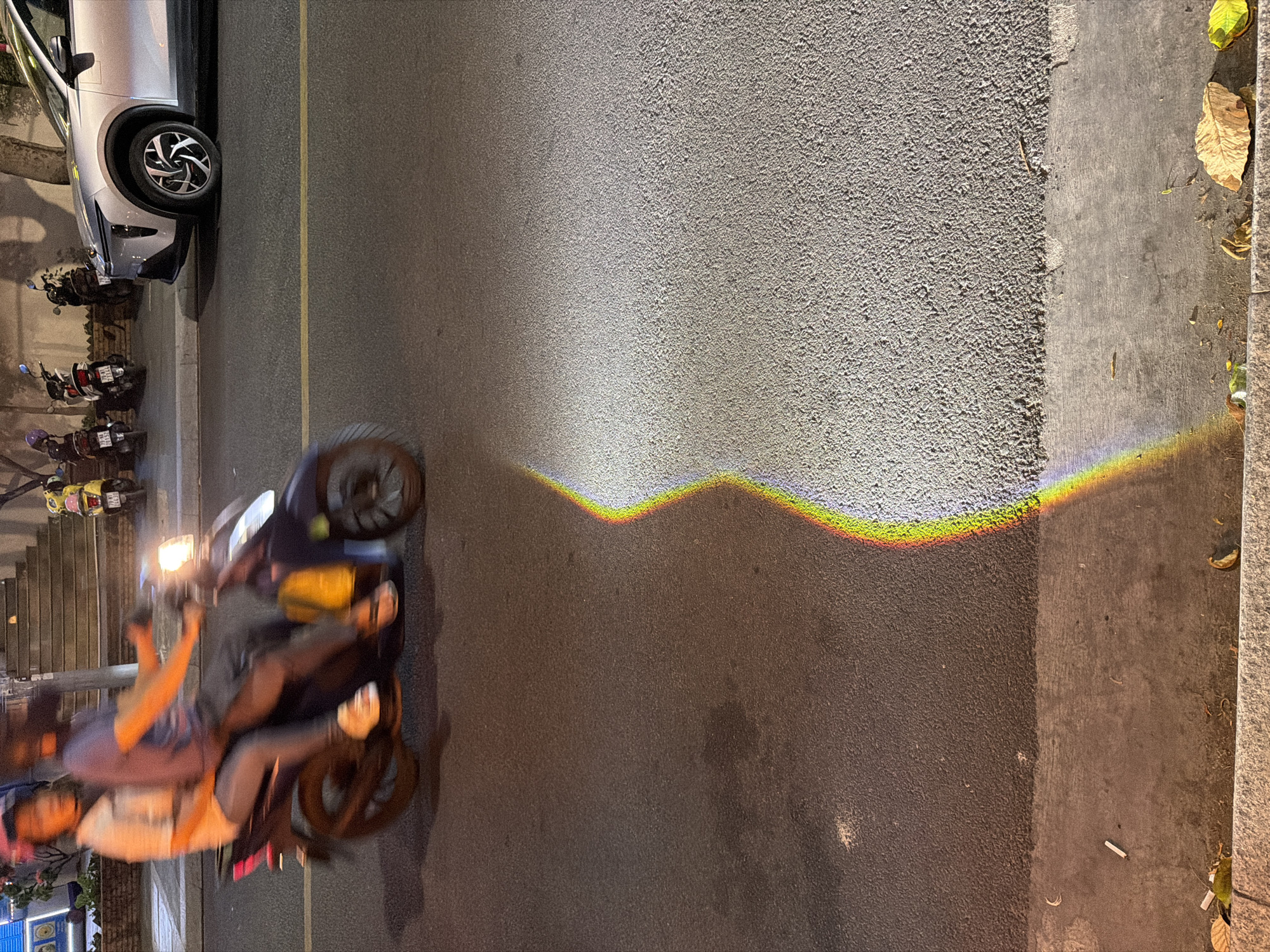 In Nha Trang Ward, Vietnam, at street level, a motorcycle with two occupants travels from left to right. The driver, wearing a purple shirt and dark helmet, is positioned at the front, while a passenger in a light-colored shirt and pink helmet sits behind. The motorcycle is in motion, evidenced by motion blur. To the right, a silver car is parked. The road surface is asphalt, with a section of gravelly material to the right. A distinct rainbow-like light refraction is visible on the road, casting colorful light streaks. The background shows buildings with stairs and several parked motorcycles. Debris, including leaves and a cigarette butt, is present on the sidewalk.