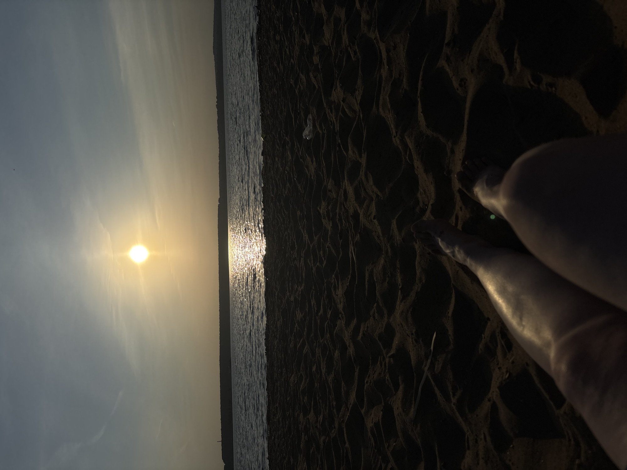 The image shows a beach at sunset, likely in Perm, Russia, based on the provided context. The sky is a gradient of blue to yellow, with the sun low on the horizon, casting a glare on the water. The foreground is dark sand, with the legs and feet of a person visible in the lower right. The water's edge is lined with a dark treeline. There's some discarded transparent object (possibly a bottle) visible in the sand. The person appears to be relaxing on the beach, observing the sunset.