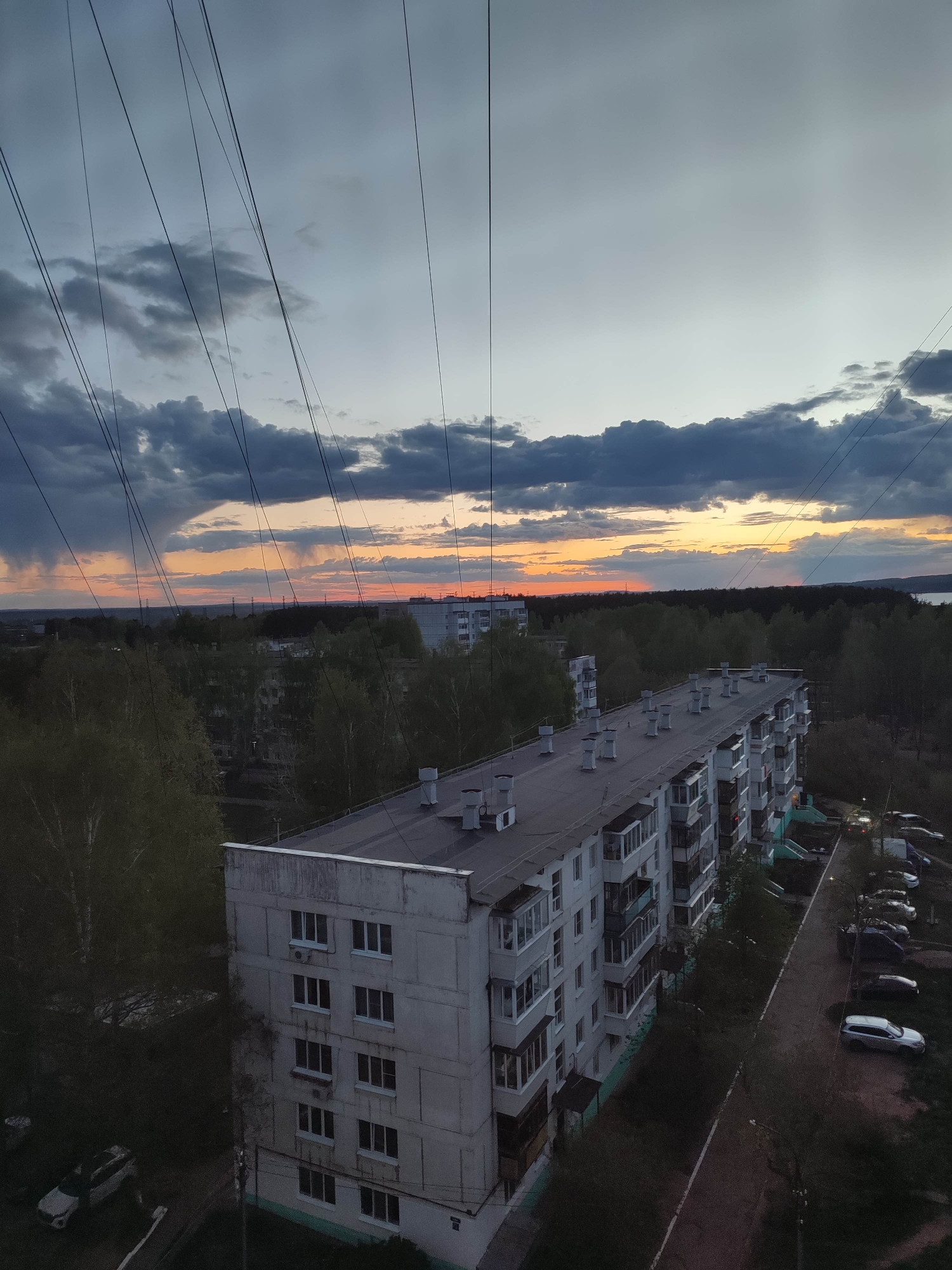 An elevated, aerial view depicts a long, multi-story apartment building in an urban area. The building has a flat roof with numerous ventilation or exhaust pipes. Balconies are visible along one side of the building. A narrow road runs alongside the building, with parked cars along it. Trees surround the building and populate the foreground. In the background, a forest line is visible with a body of water beyond it. The sky is filled with dark, stormy clouds, with the setting sun casting orange hues along the horizon. Power lines are strung across the upper portion of the image. The overall lighting suggests it is dusk or twilight.