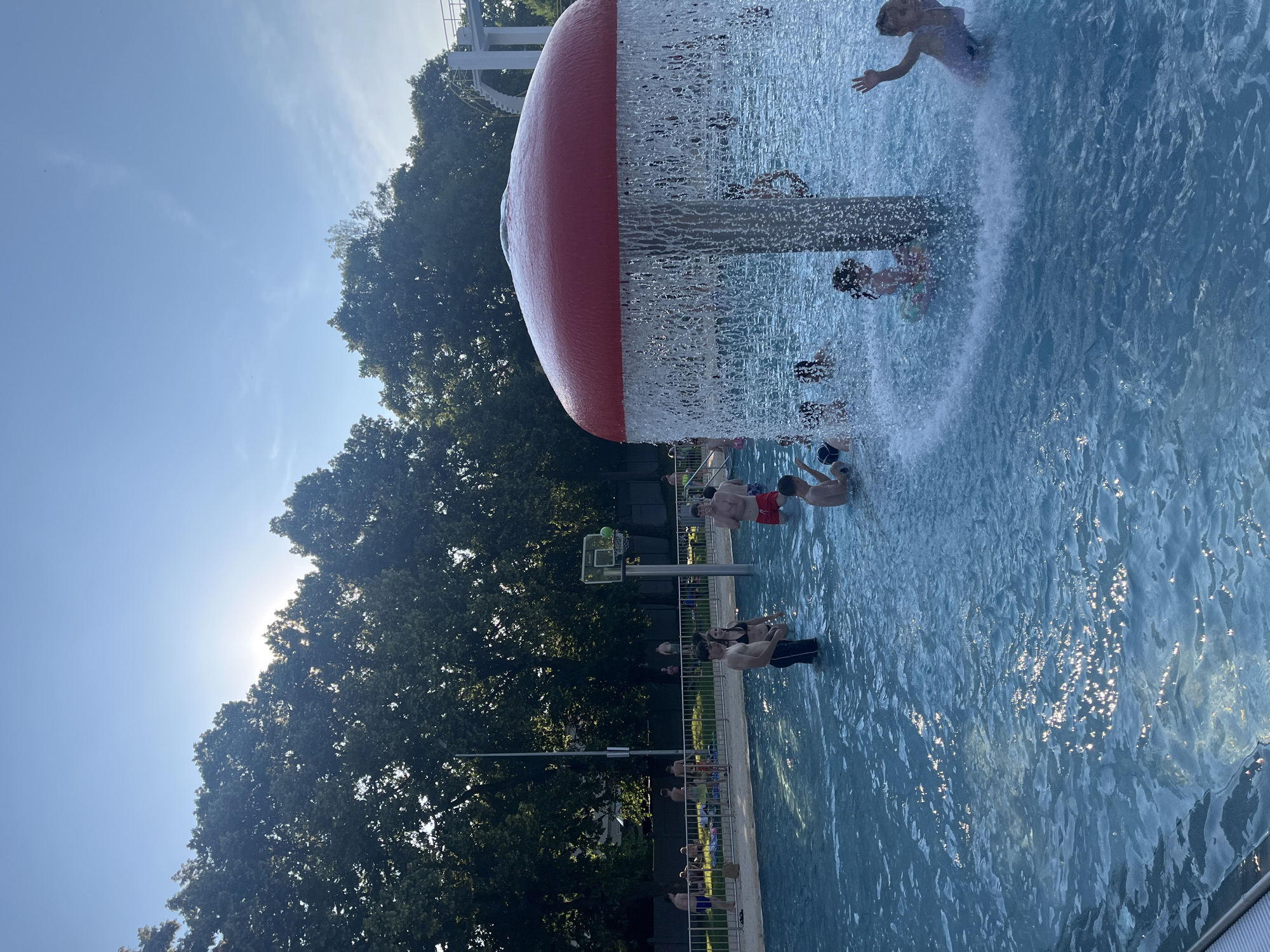 This image depicts an outdoor swimming pool area on a sunny day. In the foreground, a mushroom-shaped water feature sprays water over a group of children playing in the shallow pool. The water is clear blue and reflects the sunlight. In the mid-ground, several adults and teenagers are standing and walking in the pool, some interacting with each other. A basketball hoop is visible in the background, with a basketball in the net. Beyond the pool, lush green trees line the perimeter of the area. The sky is a clear blue with some wispy clouds. The overall scene suggests a recreational and lively atmosphere.