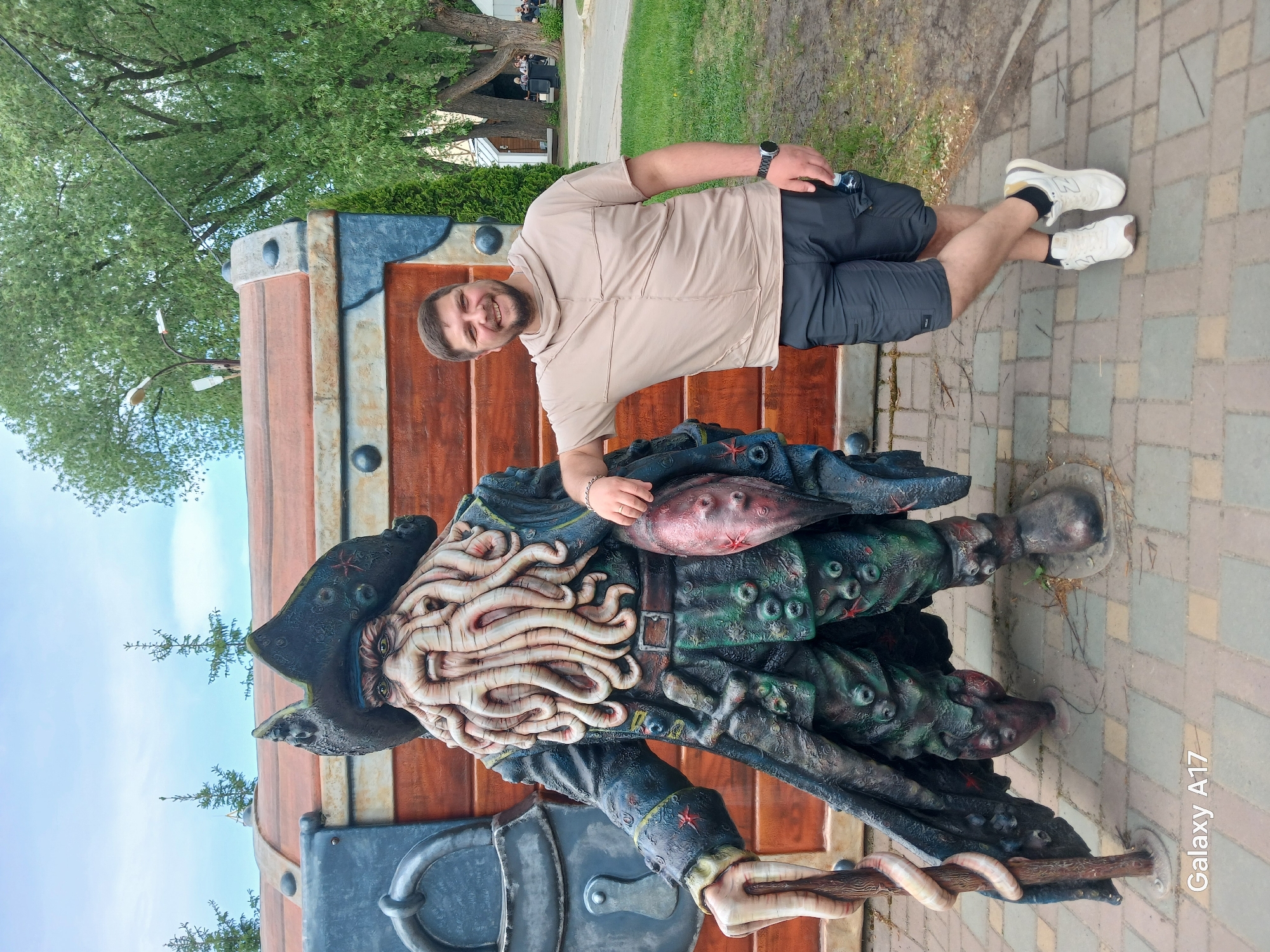 A man is posing with a statue of Davy Jones from "Pirates of the Caribbean," in Yekaterinburg, Russia. The statue is in front of a wooden chest. The man is wearing a tan shirt, dark shorts, and white sneakers. He is leaning on the statue and smiling. The statue is detailed, with tentacles for a beard, and wearing a pirate hat. The scene is outdoors, on a brick paved area. Trees and grass are visible in the background. "Galaxy A17" is written in the lower right corner.