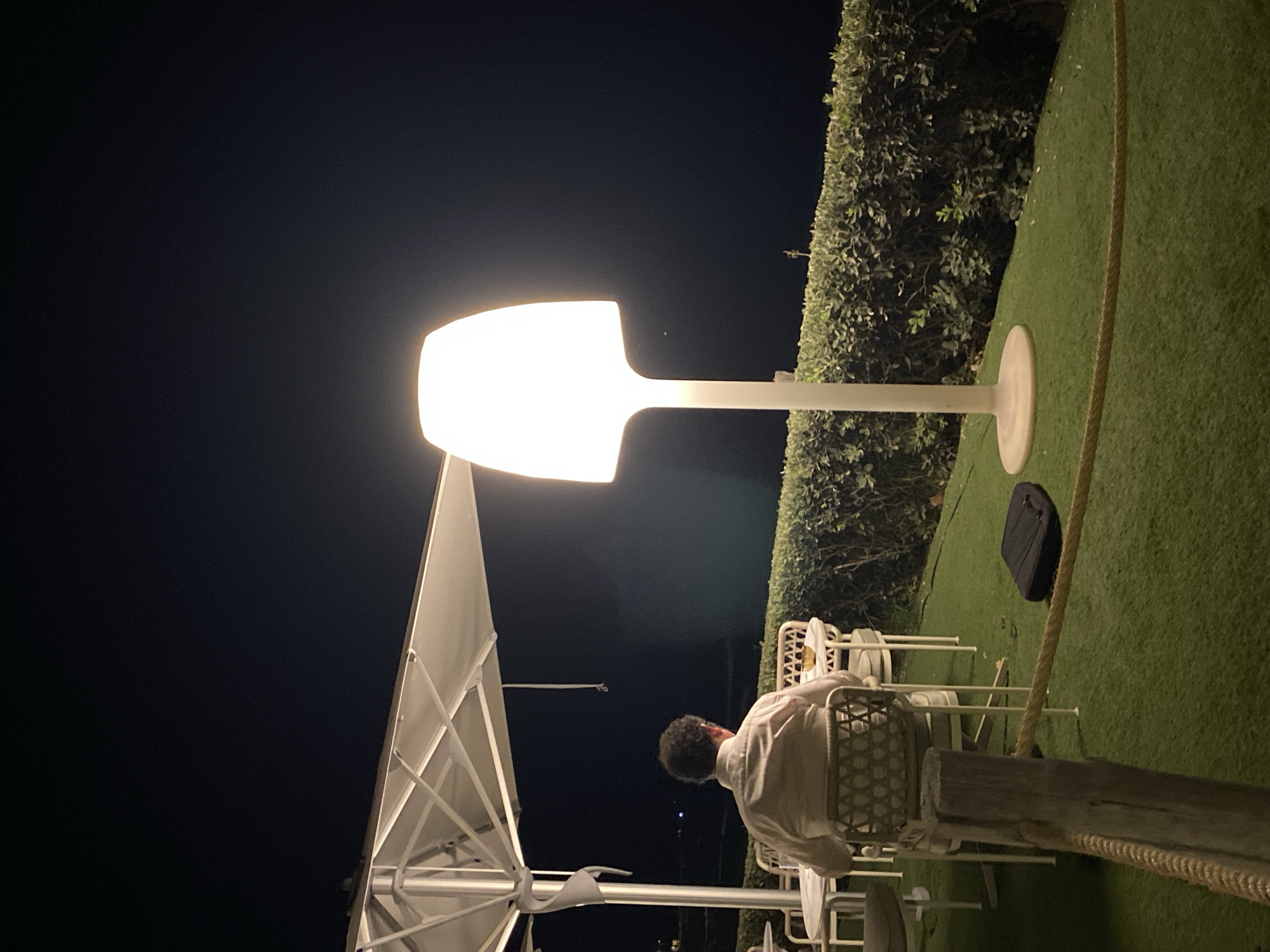 A person sits at a table under an umbrella at night on a lawn with an artificial grass surface. The person, viewed from behind, wears a light-colored long-sleeved shirt. In the foreground, a thick rope is secured to a wooden post. A large, modern, illuminated floor lamp stands to the right of the frame, casting a bright light. Behind the person and table is a well-maintained hedge. The scene appears to be an outdoor seating area, possibly a restaurant or bar, in Estepona, Spain. The sky is dark, indicating nighttime.
