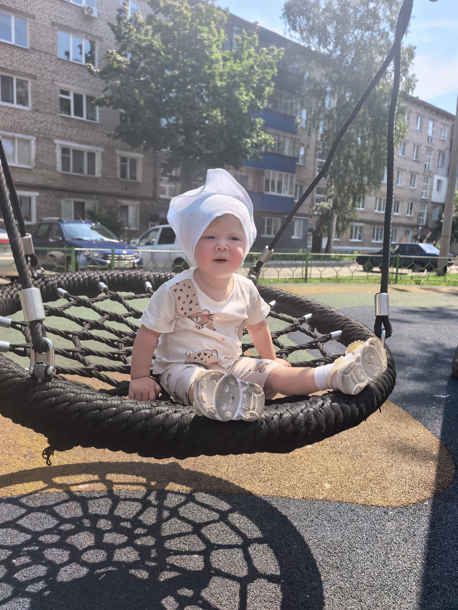 A young child sits on a round, black net swing on a sunny day in a playground in Sterlitamak, Russia. The child is wearing a light-colored t-shirt and shorts with animal prints, and a white headscarf. The swing is suspended by thick black ropes. In the background, there are multi-story brick apartment buildings and trees. The ground of the playground is made of rubberized material in yellow and dark gray. The child is looking towards the camera with a slight smile. The sun casts a patterned shadow of the swing onto the ground.