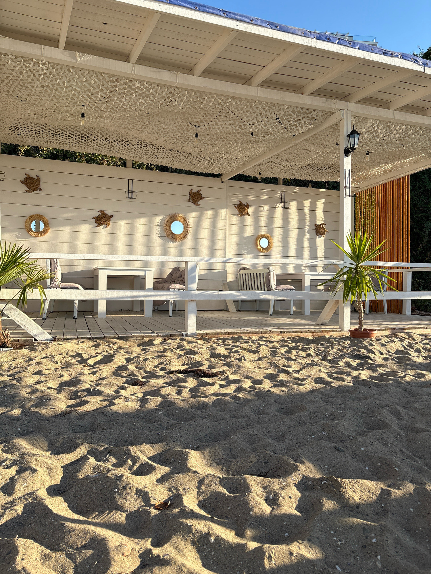 This image depicts an outdoor seating area on a sandy beach, likely in Sveti Vlas, Bulgaria. The structure is a white wooden gazebo with a slatted roof covered by a white camouflage net. String lights are suspended from the ceiling. The back wall of the gazebo is made of white wooden planks, decorated with four turtle-shaped wall hangings and three round, woven mirrors with sunburst patterns. In front of the wall, a white wooden bench with cushions and a white table are visible, accompanied by chairs. To the left, a palm frond extends into the frame, and to the right, a potted palm tree stands near a wooden fence. The foreground is dominated by rippled sand, with shadows cast by the gazebo and nearby foliage. The sky above is clear and blue. The scene appears to be set during daylight hours, with the sun casting long shadows. There are no people visible in the image.