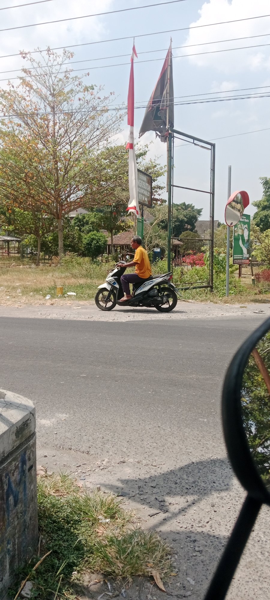 A man rides a white scooter along a paved road. He is wearing a yellow t-shirt and dark pants. The scooter is moving from left to right. In the background, there are trees, some buildings with red tile roofs, and two flags on poles. One flag is red and white, and the other is dark. A convex mirror is mounted on a pole to the right of the flags. The ground is dry and there is some grass along the side of the road. The sky is visible above the trees and flags. The image appears to be taken in daylight. The scene suggests a rural or semi-rural area in Maguwoharjo, Indonesia, indicated by the presence of flags and architecture.