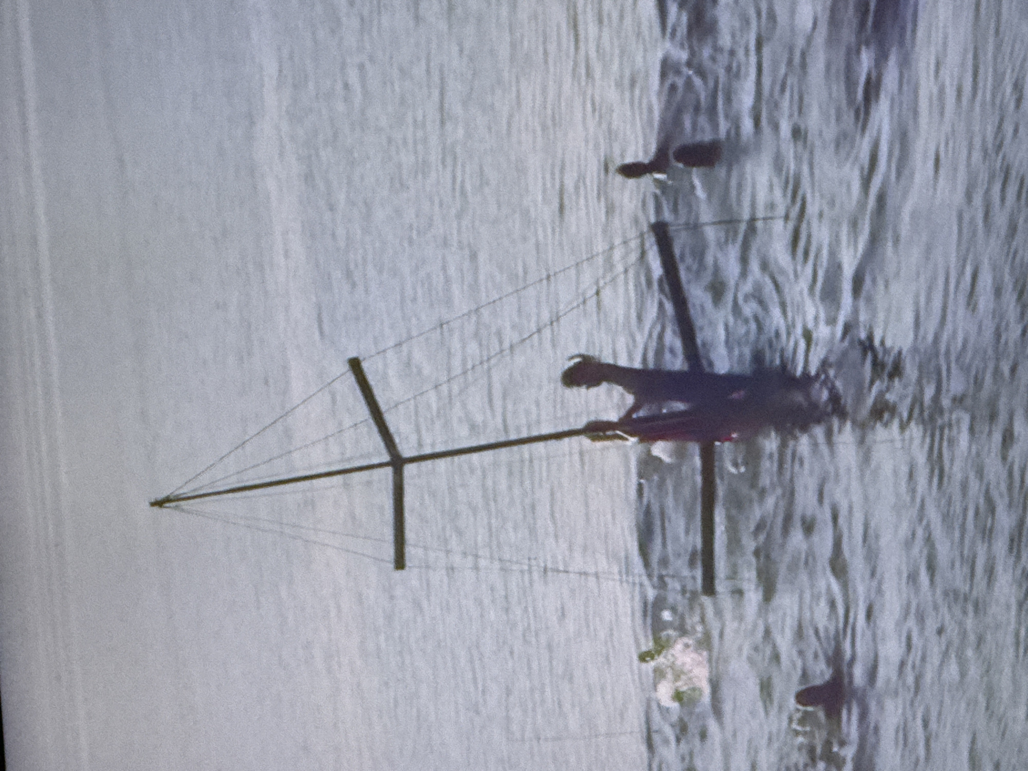 In the city of Unknown, Unknown, a group of individuals are present in a body of water. Several people are partially submerged in the water, with some appearing to swim or float. In the center of the image, a rudimentary boat-like structure with a mast and rigging is visible, with at least two individuals on or near it. One individual is standing and appears to be holding onto the structure. Another individual is also on or near the boat. To the left of the boat, a person is in the water with arms raised. To the right of the boat, another person is visible in the water. In the distance, on the right side of the image, another person is visible in the water, and further to the right, another individual's head is above the water. The water is calm with some ripples. The sky is overcast, and the lighting suggests it is daytime. The scene depicts people interacting with a simple watercraft in a body of water.