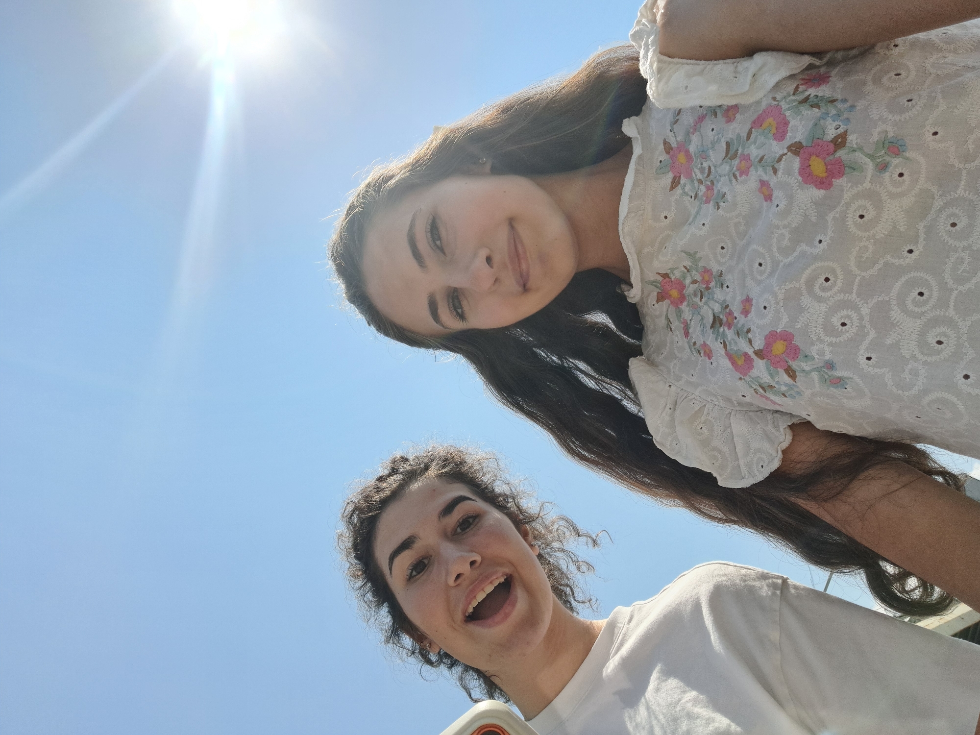 Two young women are posing for a selfie against a bright blue sky with the sun glaring in the upper right corner. The woman on the left has curly brown hair and is wearing a white t-shirt. She is smiling with her mouth open. The woman on the right has long, dark brown hair and is wearing a white top with floral embroidery. She is looking towards the camera with a slight smile. The camera is positioned below them, looking up. The image appears to have been taken outdoors on a sunny day.