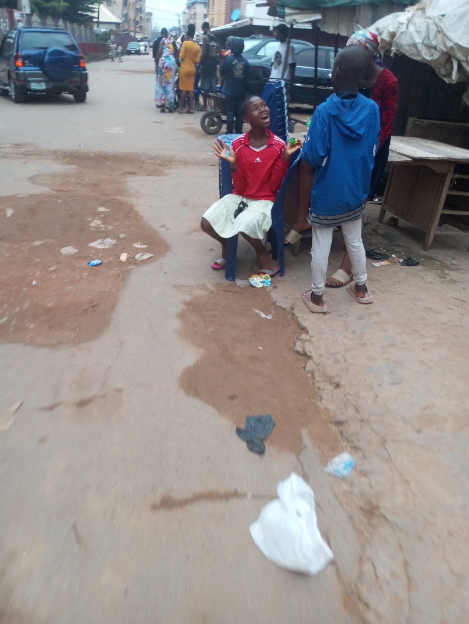 In the city of Unknown, Unknown, on a dirt road, a blue SUV is parked on the left side of the road. In the middle of the road, two children are standing. The child on the left is wearing a red shirt and white shorts. The child on the right is wearing a blue hoodie and striped pants. They appear to be interacting. In the background, several other people are standing around, and a wooden table is visible. There are also some discarded items on the ground, including plastic bags and wrappers. The overall scene appears to be a bustling street in a residential area.