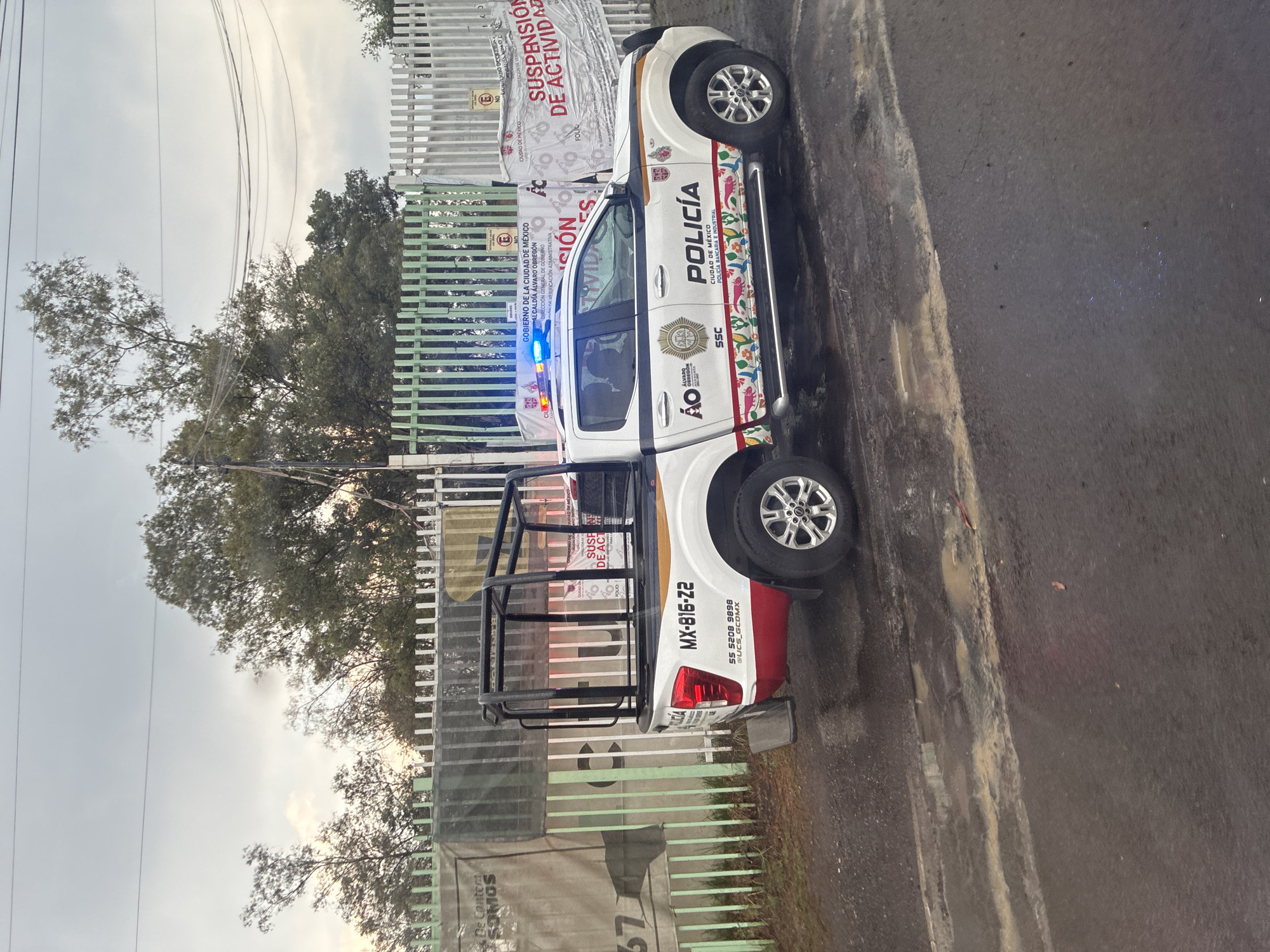 The image depicts a white police pickup truck parked on a street, presumably in Mexico City, Mexico. The truck has "POLICIA" printed on its side, along with the seal of the city and the initials SSC (Secretaria de Seguridad Ciudadana). The vehicle is adorned with colorful patterns running along the lower side panels. The license plate number is MX-816-22. The back of the vehicle is red.
In the background, a green fence is visible, with signage attached indicating "GOBIERNO DE LA CIUDAD DE MEXICO" and stating "SUSPENSIÓN DE ACTIVIDAD". Trees and overcast sky are also visible in the background. The ground is paved and wet.