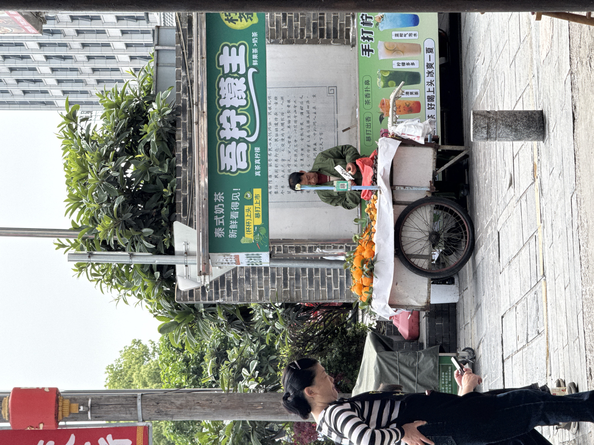 The image shows a street scene in Zhangjiajie, China. A vendor is selling fruit and tea from a cart. The cart has oranges, loquats, and beverages. A woman in a black and white striped shirt is looking at her phone in the foreground. A sign behind the vendor advertises "Lemon King" tea and other drinks, with Chinese characters and images of various beverages. The sign also has a QR code. A multi-story building is visible in the background on the left side of the image. The day is sunny.