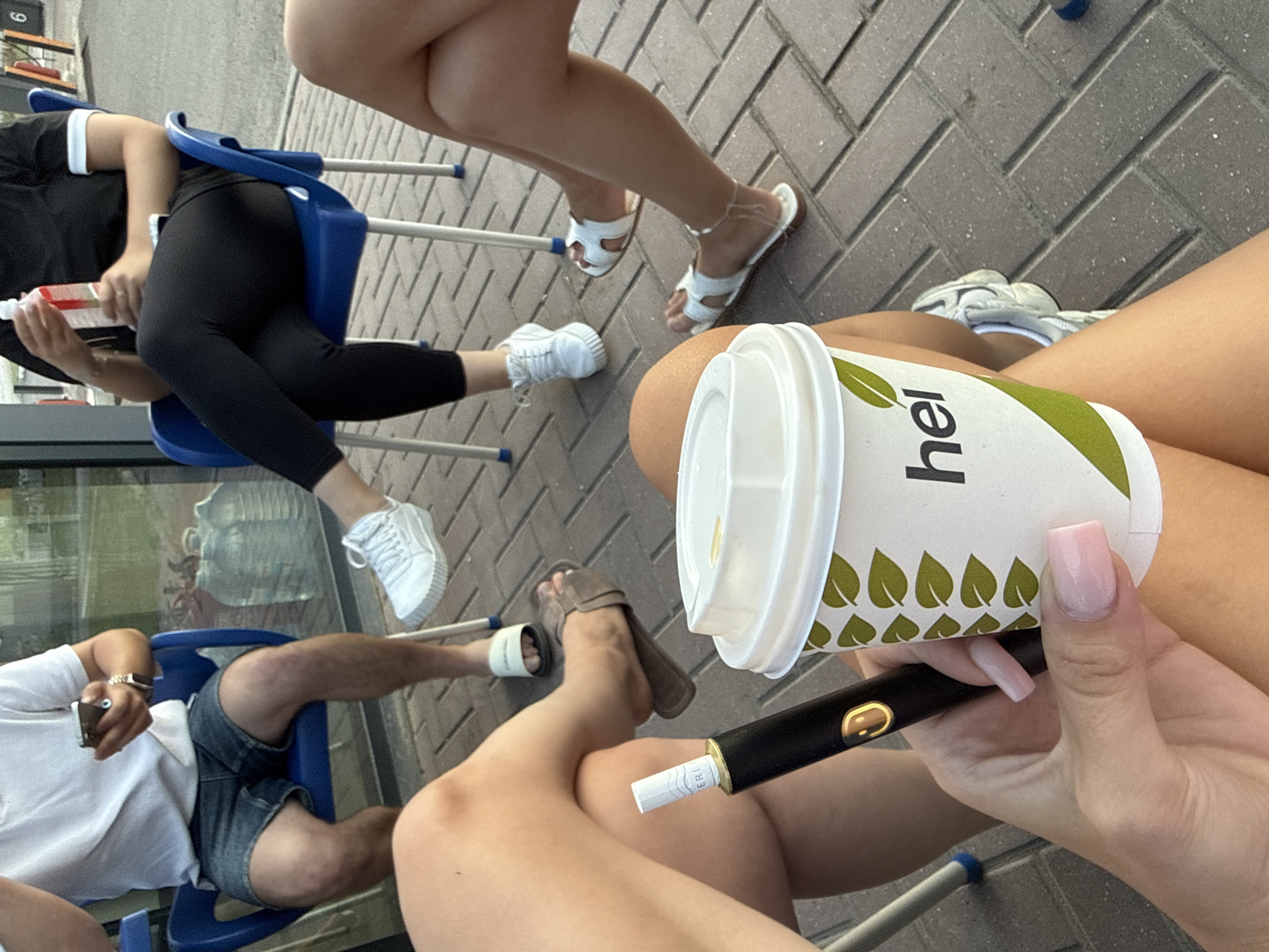 The image depicts a group of individuals seated outdoors on a paved area, likely a public space or patio. In the foreground, a person's hand with light pink square-shaped nails holds a black vaping device and a white paper cup with green leaf accents and the text "hei" printed on it. The cup appears to contain a beverage.
Other individuals are visible in the background, seated in blue plastic chairs. One person, wearing a white t-shirt and denim shorts, holds a smartphone. Another person is dressed in black leggings and a black t-shirt with white trim on the collar. A third person's legs are visible, wearing white sandals. The environment suggests an urban setting with a building facade and large water containers in the background, hinting at a commercial area such as a cafe or restaurant. There are no specific visual cues that place this scene in Brătianovca, Moldova.
