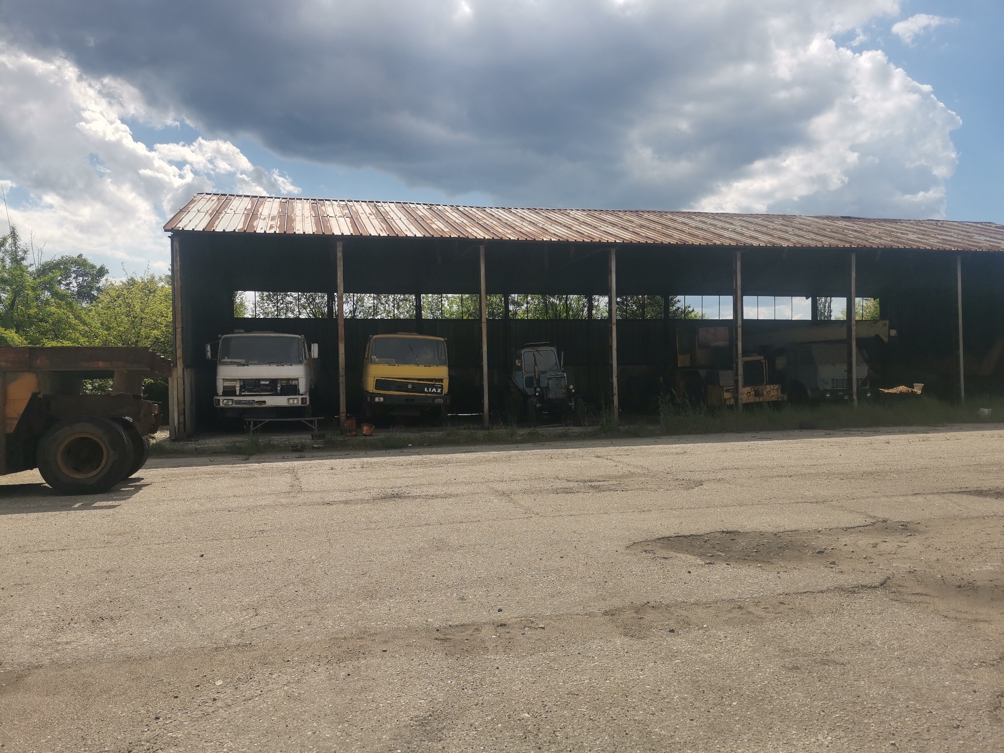 The image depicts a large, open-sided metal structure housing several vehicles. The structure appears to be a storage shed or garage, with a corrugated metal roof supported by pillars. Inside, there are multiple trucks and construction vehicles of varying sizes and colors. A white truck is on the left, a yellow truck is next to it, and a blue truck is behind them.
The structure is located on a paved surface with some cracks and disrepair. Vegetation is visible behind the structure. The sky is visible overhead with scattered clouds.
The picture was taken in Zhivkovo, Bulgaria.